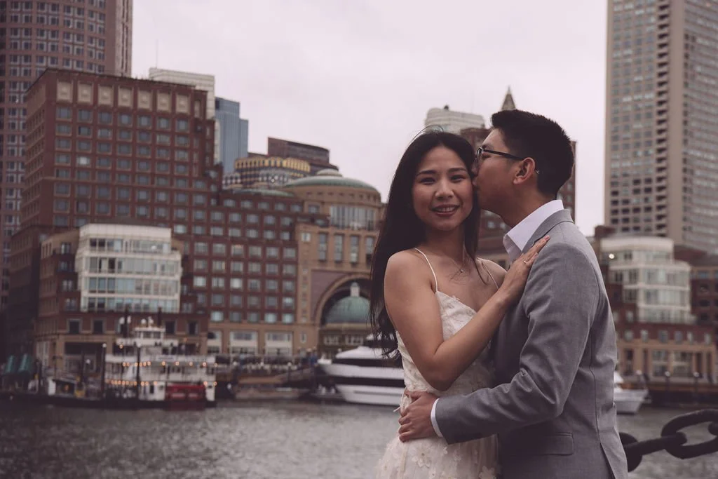 Justin kissing his future bride's cheek at Fan Pier Park with the Boston skyline in the background during their spring engagement session. Photography by Mike Indi.
