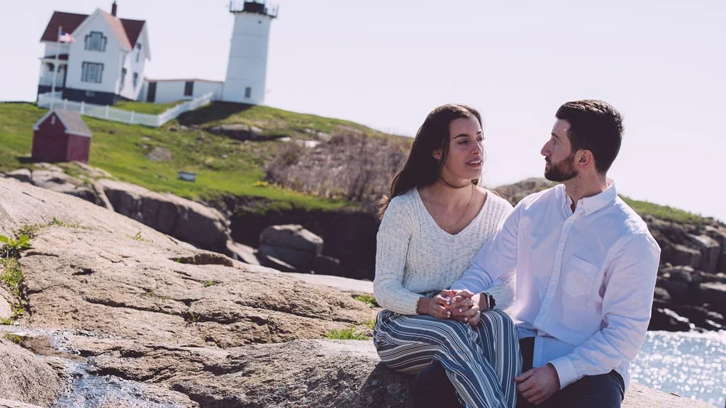 Carol sitting and holding her new fiancé's hand with tears of joy in her eyes during their proposal and engagement session at Nubble Lighthouse in York, ME. Photography by Mike Indi.