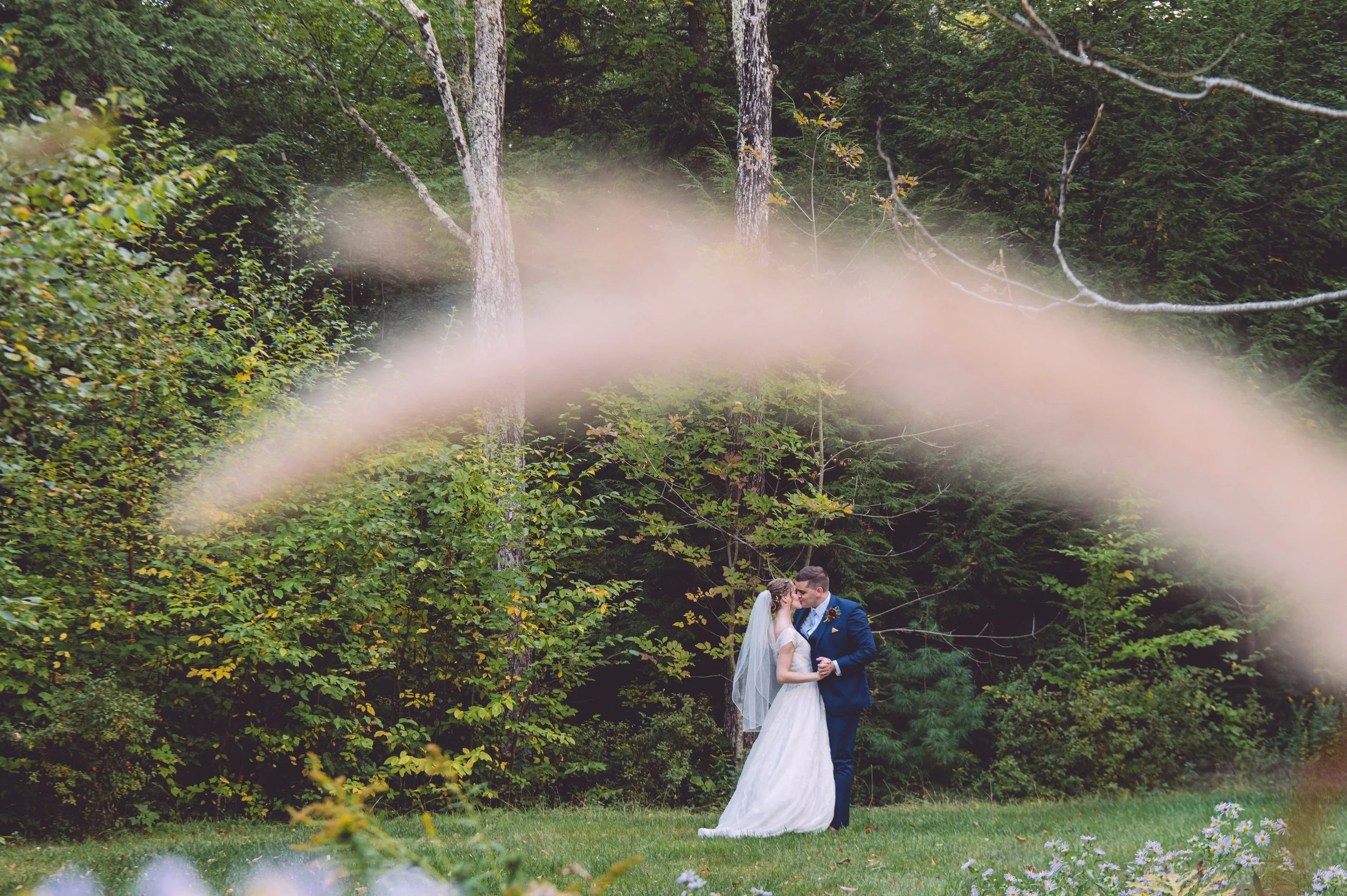 A gorgeous bride and groom sharing a passionate kiss in a field surround by wild flowers during their wedding at Locke Falls Farm in Tamworth, New Hampshire - Photography by Mike Indi