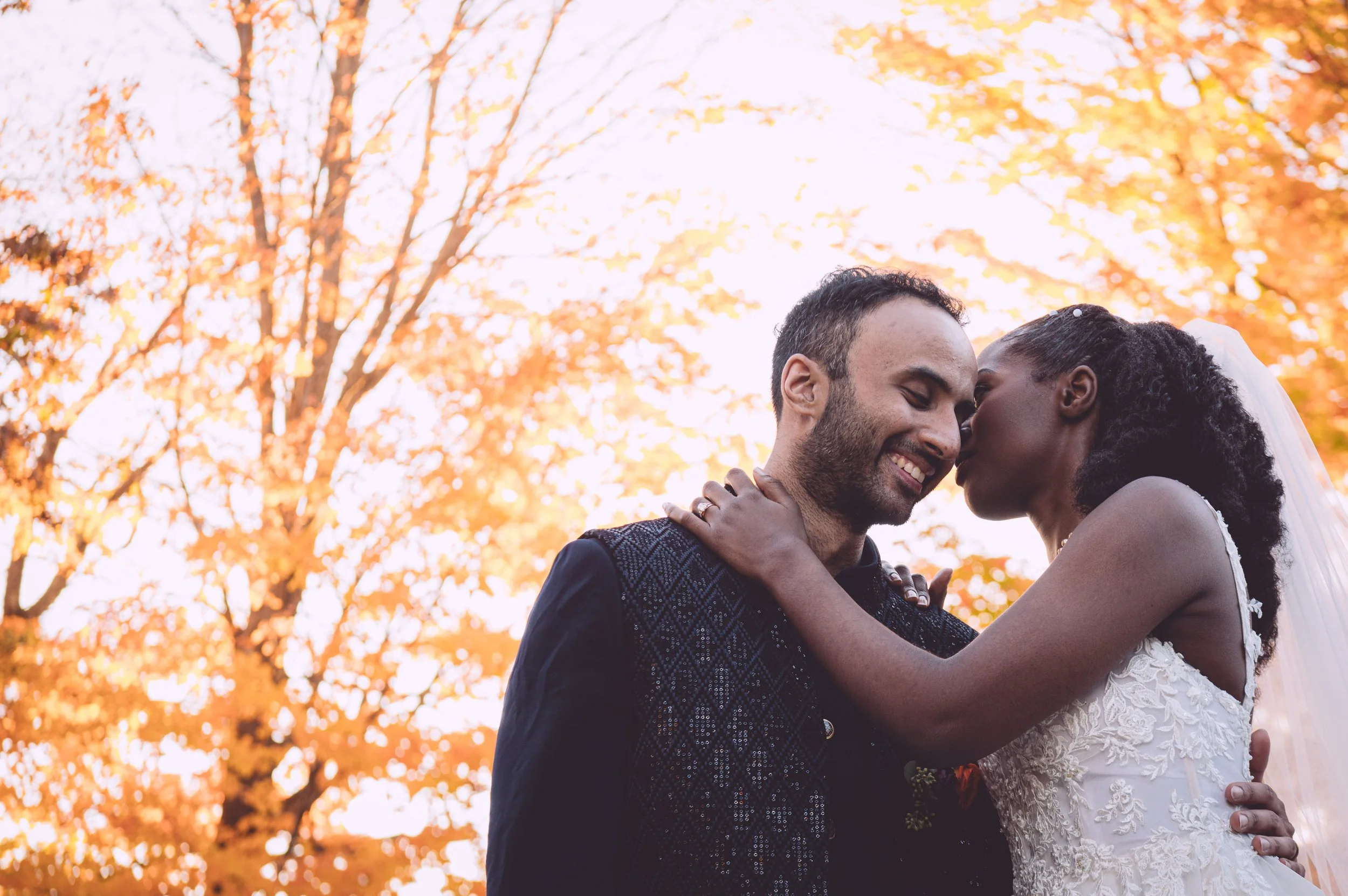 A bride and groom sharing a tender moment surround by bright orange fall foliage during their autumn wedding at Searles Chapel in Windham, NH. Wedding photography by Mike Indi.