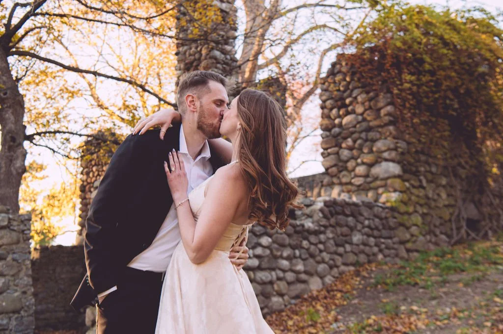A bride and groom sharing a passionate kiss beside the stone tower ruins at Bancroft's Castle in Groton, Massachusetts during a fall engagement session — photography by Mike Indi