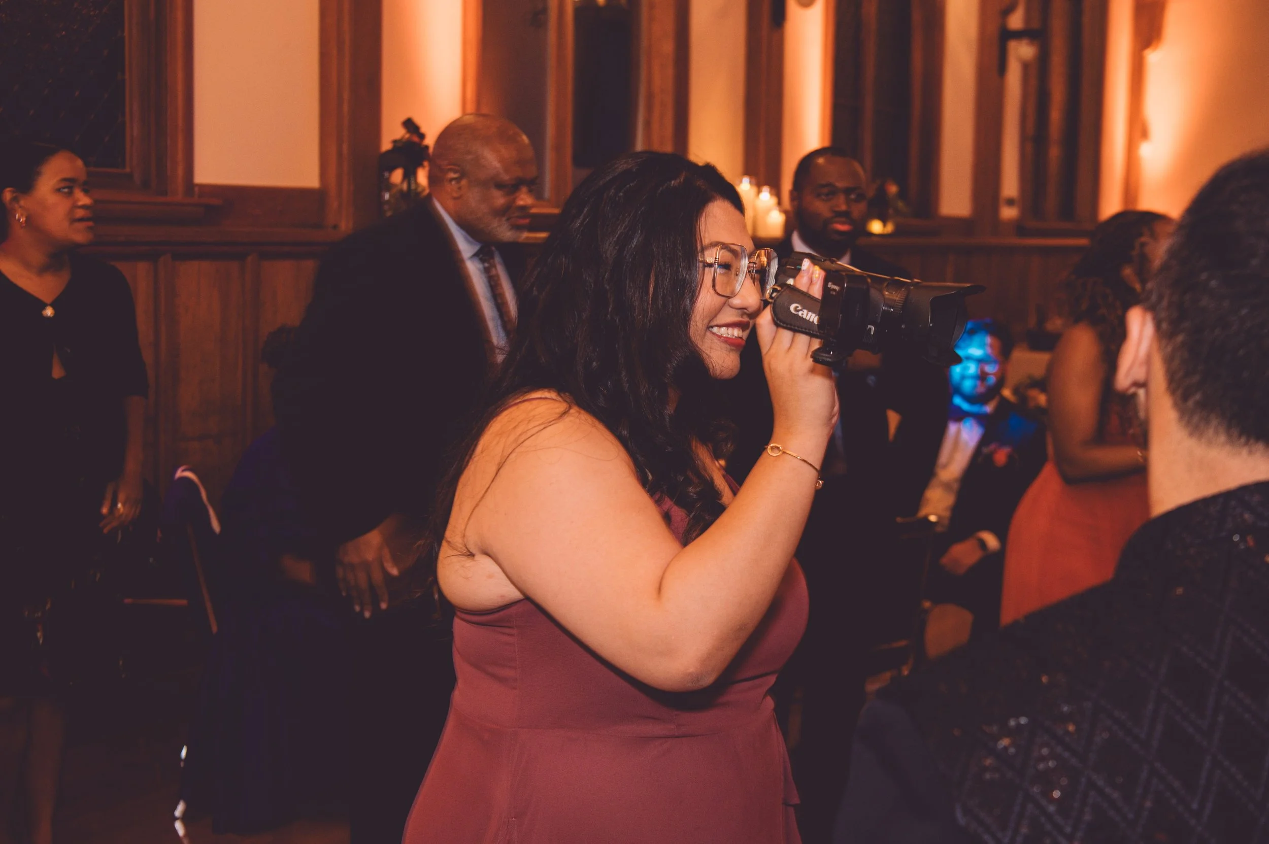 A maid of honor smiling while holding a vintage video camera to record a bride and groom dancing during their wedding reception at Searles Chapel in Windham, NH. Wedding photography by Mike Indi.