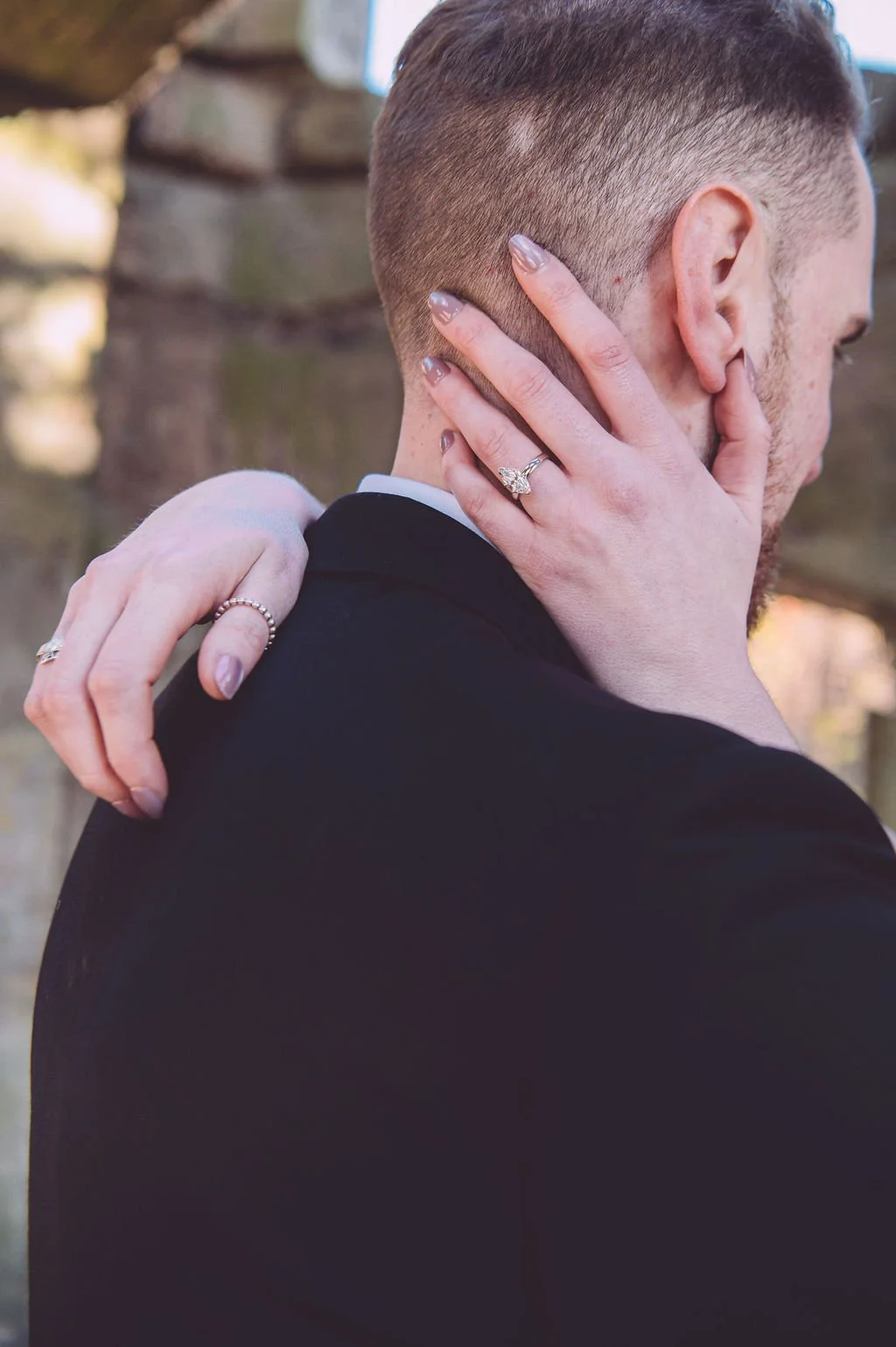 A close-up of a bride's hand featuring her wedding ring while her hand is gently caressing the back of her groom's head during their engagement session at Bancroft's Castle in Groton, MA. Engagement photography by Mike Indi.