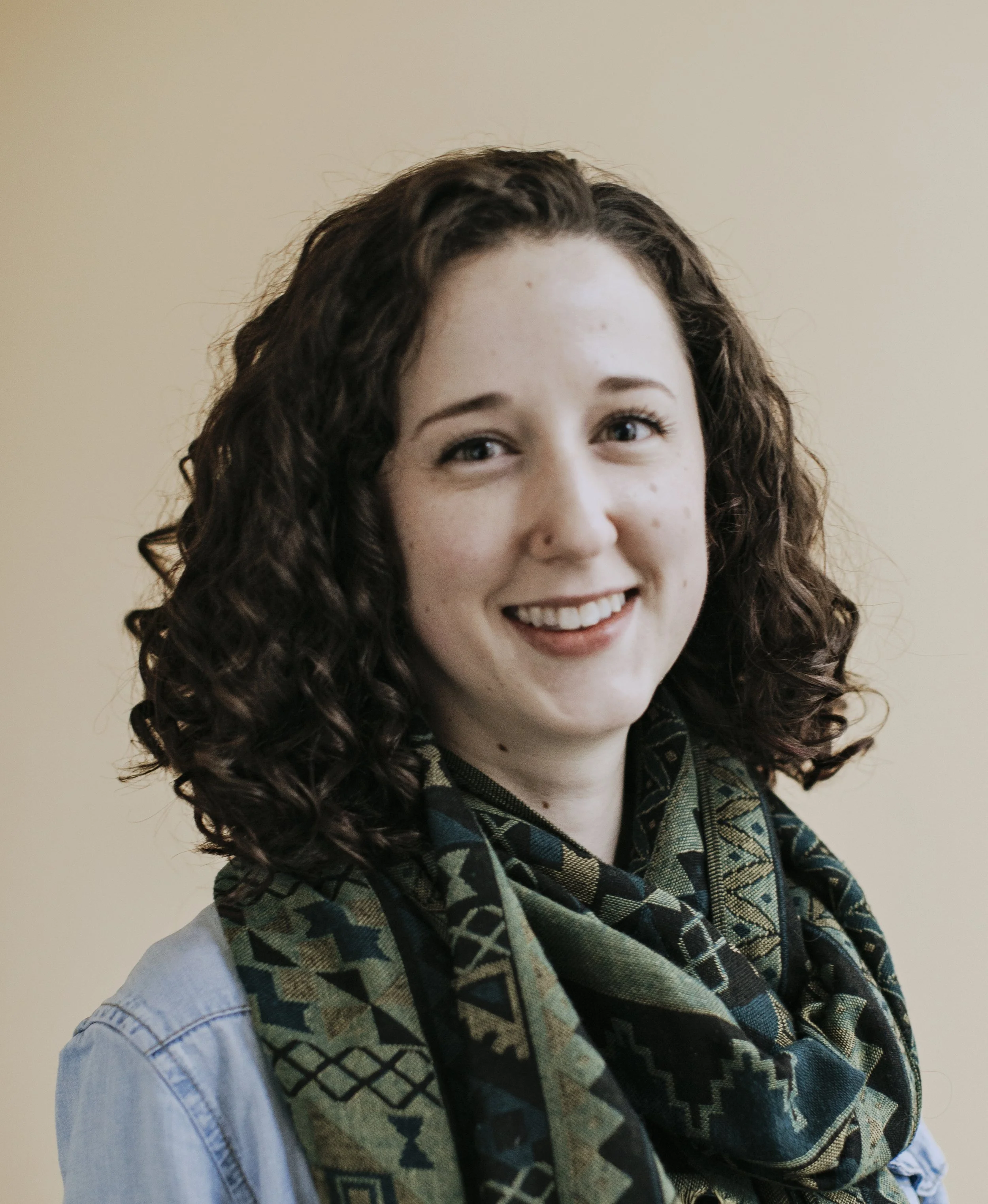A woman with curly brown hair, blue eyes, and light skin, smiling, wearing a light denim jacket and a patterned scarf, standing against a beige background.