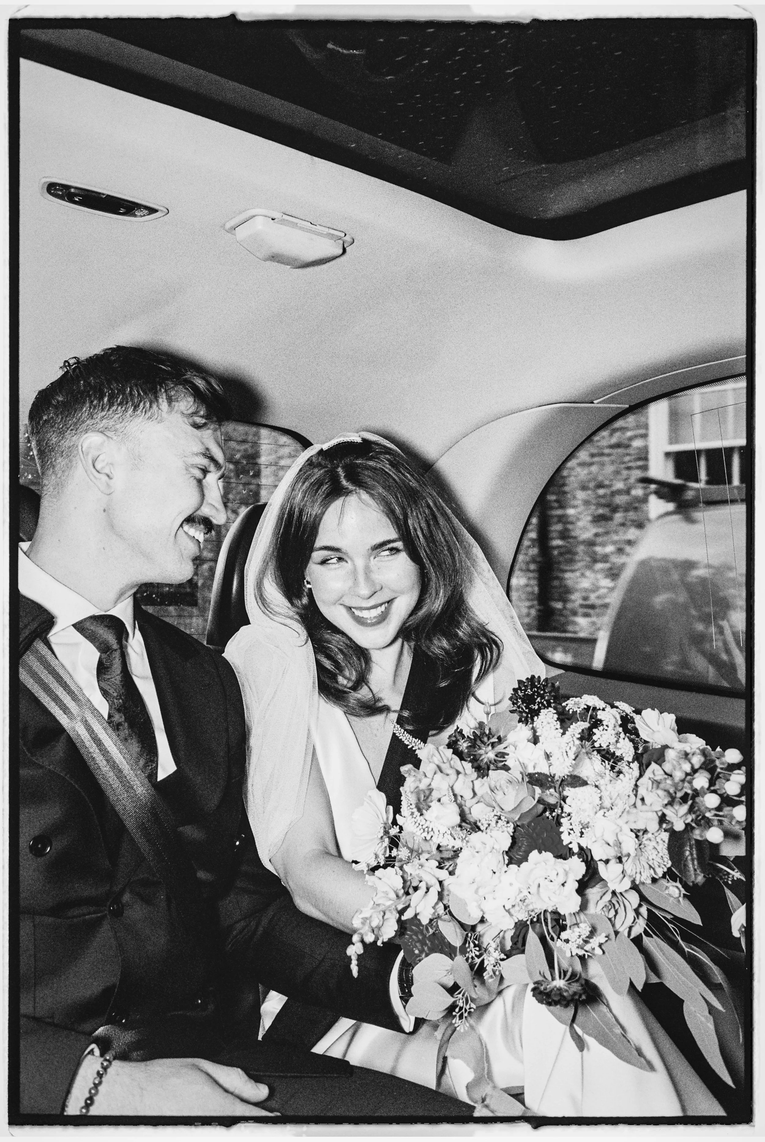 A black-and-white photo of a newlywed couple sitting in a car, smiling and laughing. The bride is holding a bouquet of flowers and wearing a veil, while the groom is dressed in a suit and tie.