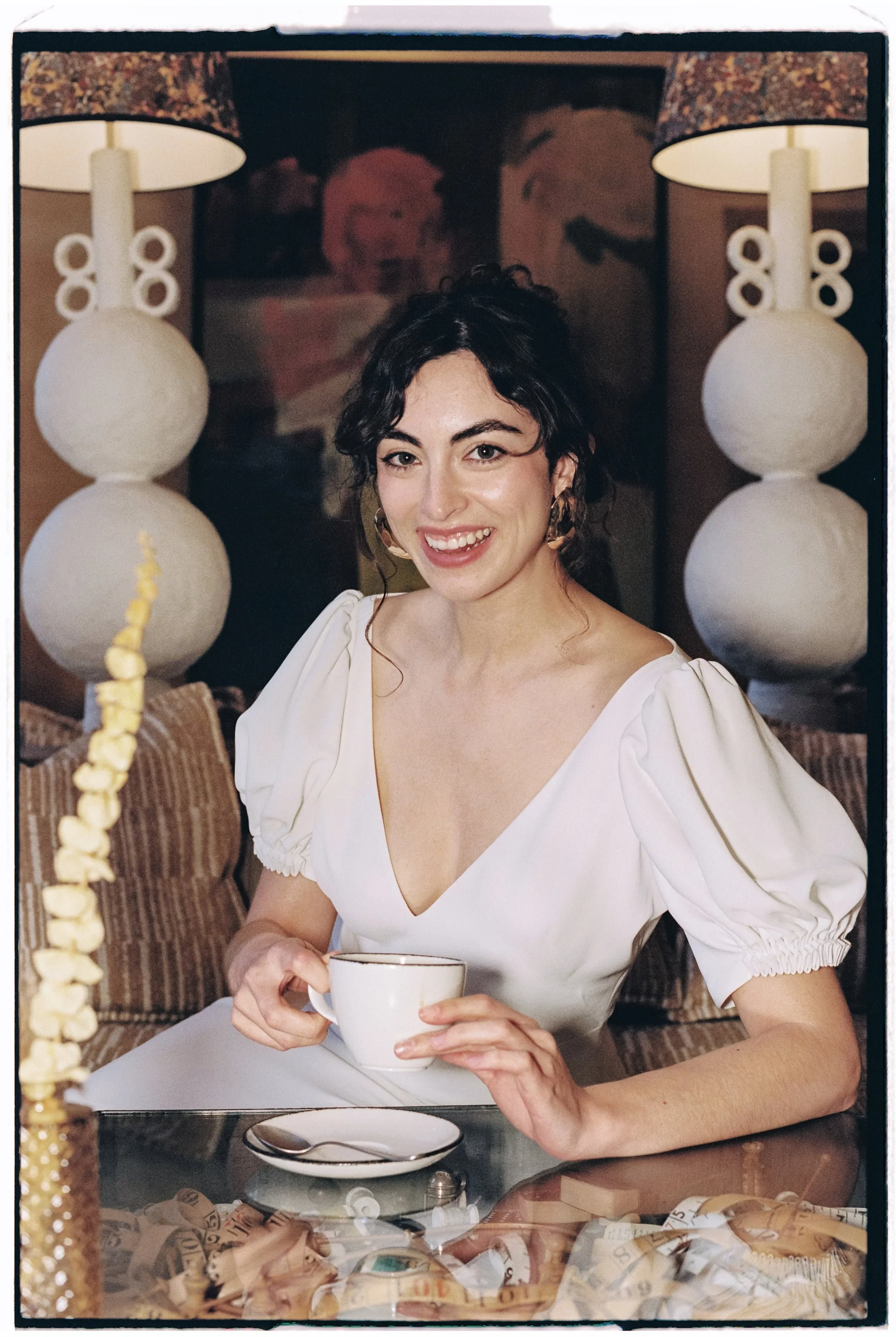 A woman with dark curly hair, wearing a white dress with puffed sleeves, is sitting at a table holding a teacup. She is smiling and looking at the camera. There are two large white lamps and a painting of cartoon characters in the background.
