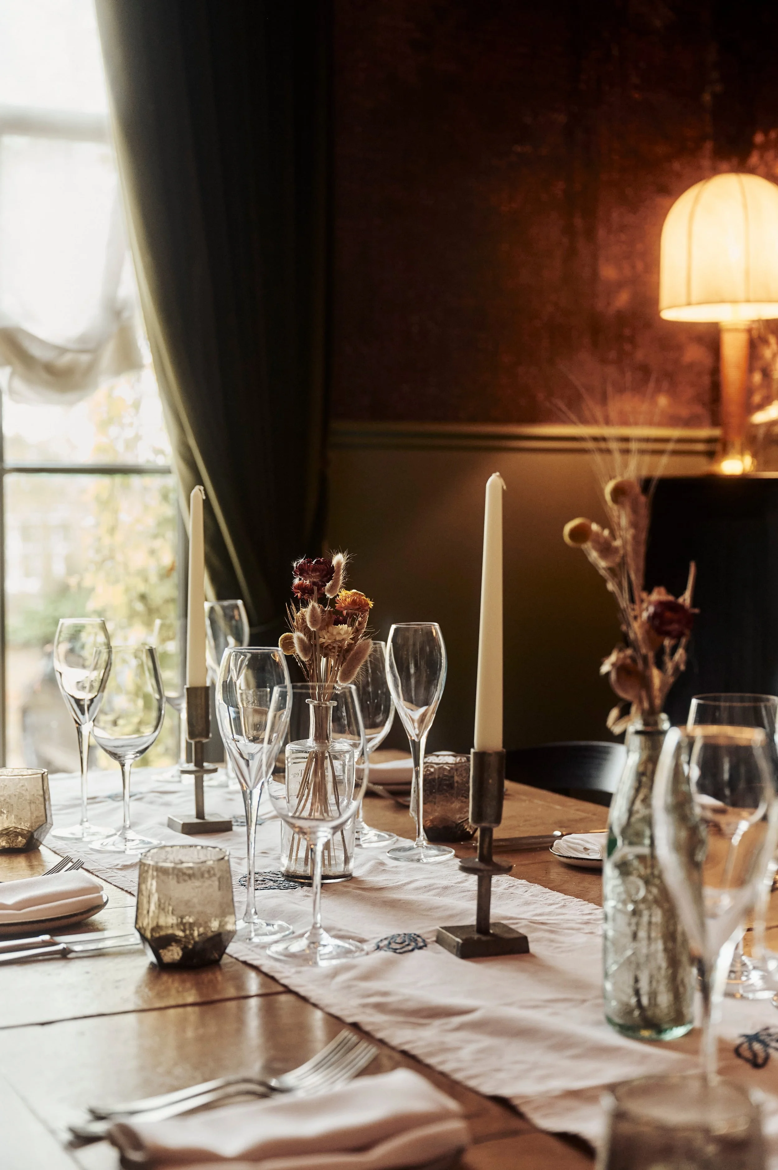 A elegantly set dining table with candles, glassware, dried floral arrangements, and a white table runner in a warmly lit room.