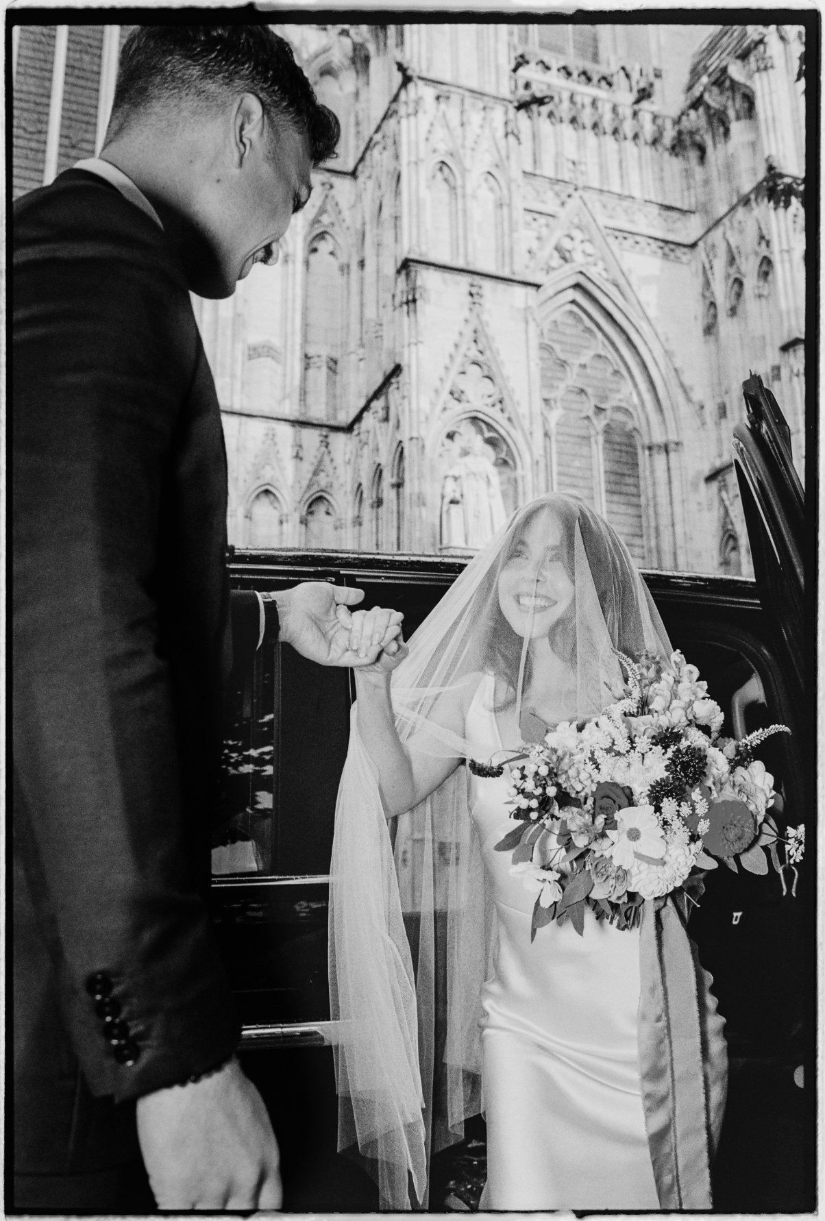A bride smiling while holding a bouquet of flowers, inside a car with a veil over her face, as a groom in a suit holds her hand outside the car with a historic cathedral in the background.