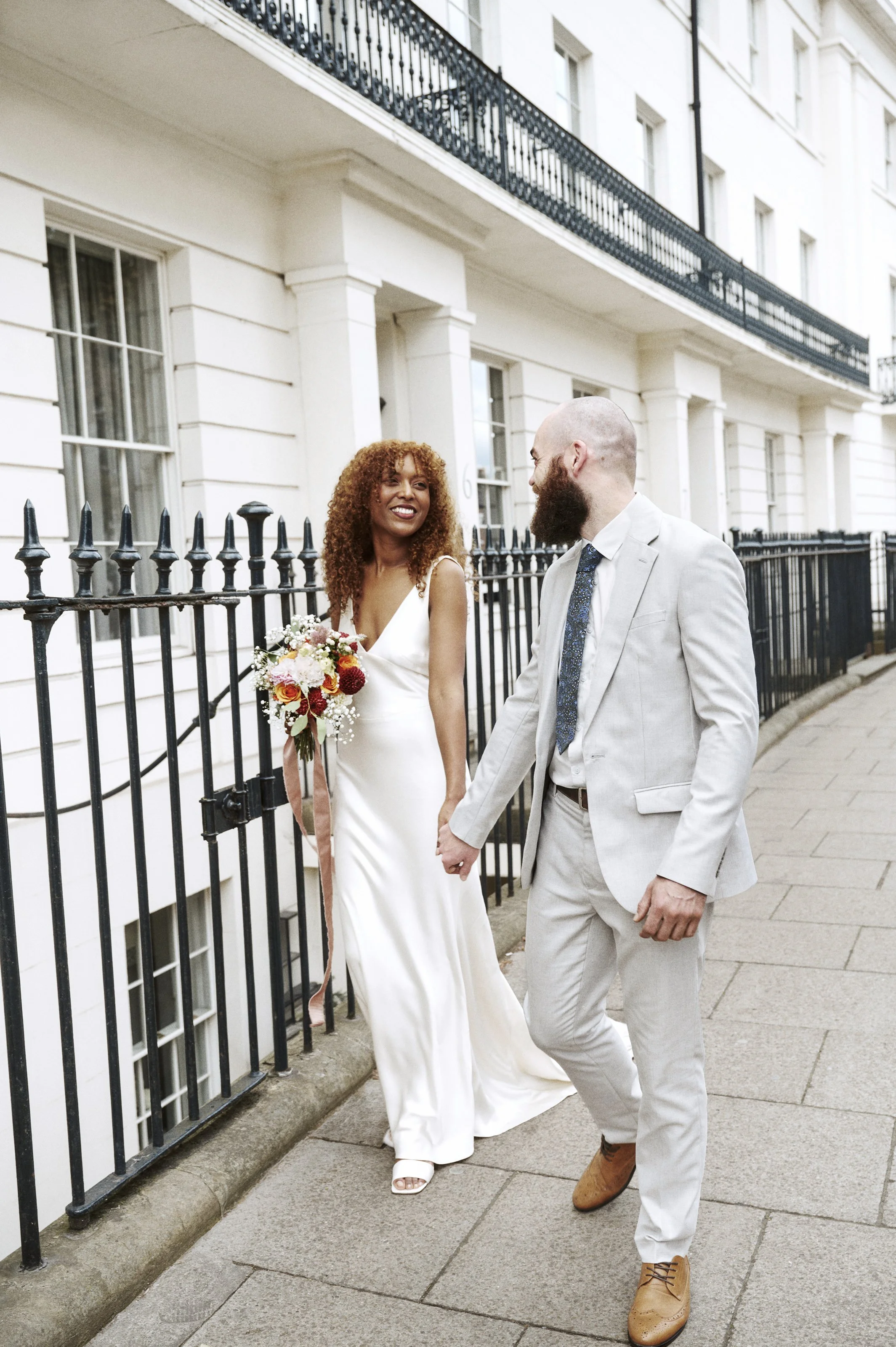 A couple, dressed in wedding attire, holding hands and smiling at each other on a city sidewalk beside white buildings with black iron fences.