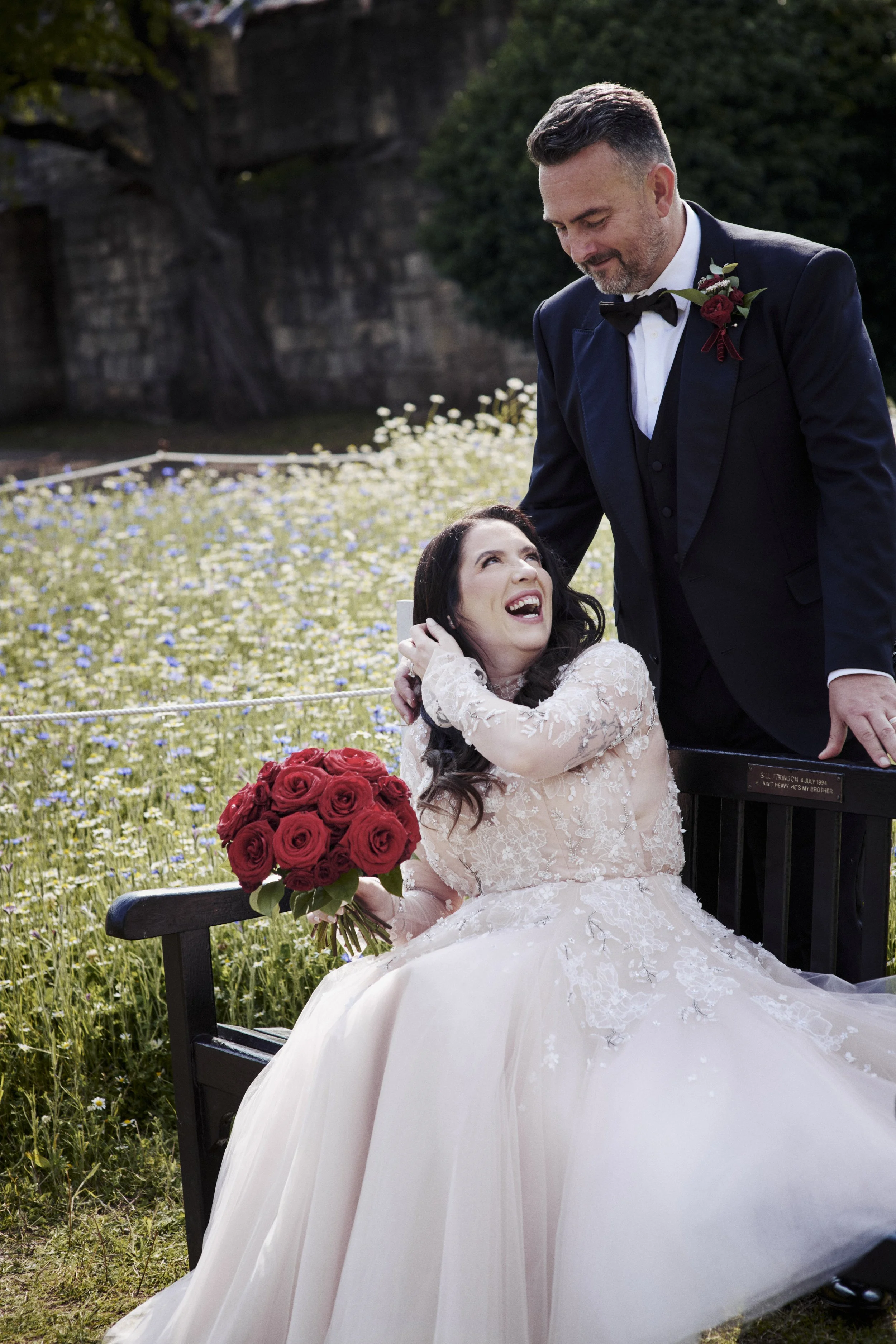 A bride in a white lace wedding gown sitting on a black bench, holding a bouquet of red roses, looking up and smiling as a groom in a black tuxedo bends over her smiling, in a field of flowers with trees in the background.