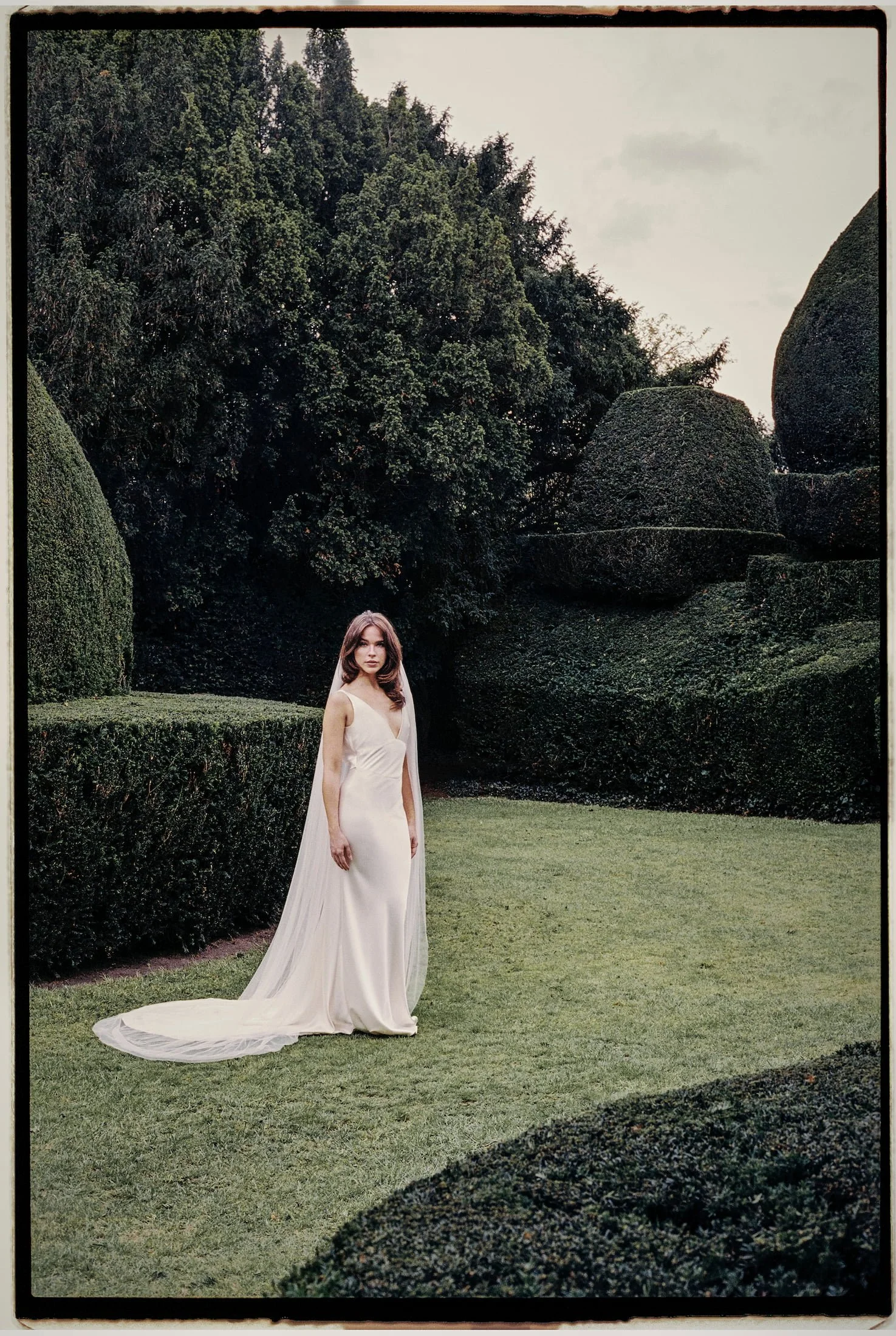 A woman in a long white wedding dress and veil walking in a garden with manicured bushes and trees.