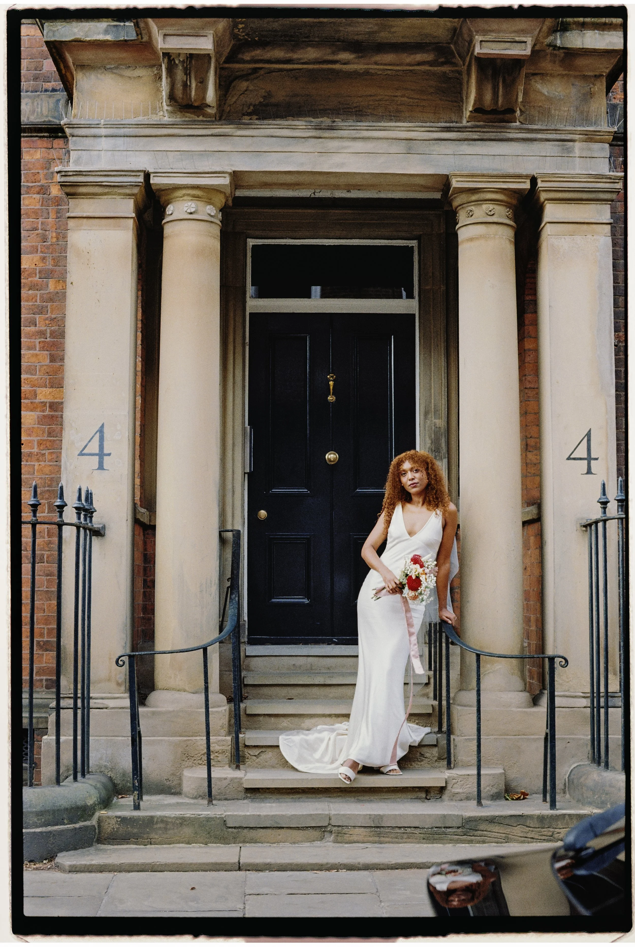 A woman in a white wedding dress holding a bouquet standing on the steps of a black front door with the number 4 on each side, flanked by stone columns and black iron railings.