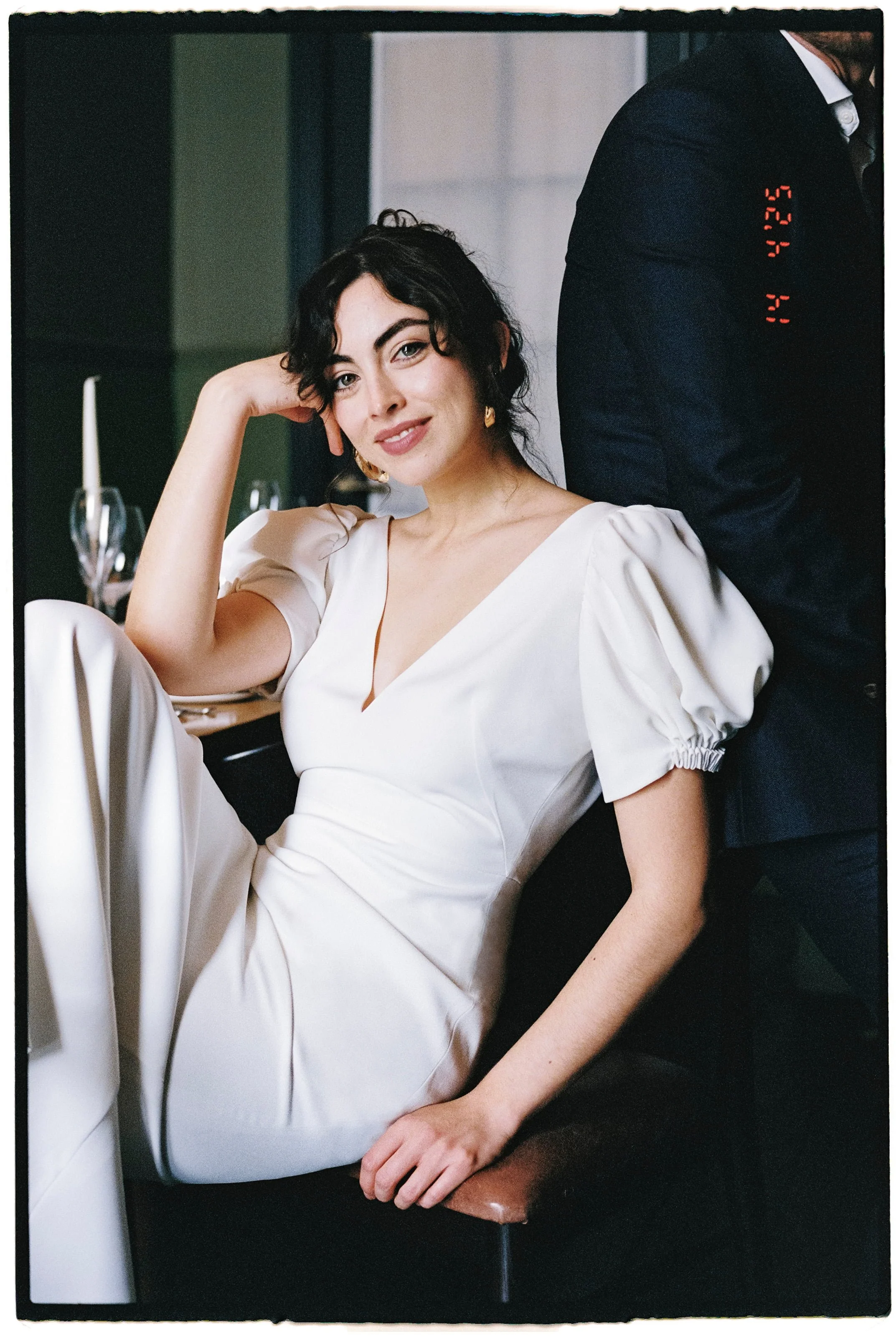 A woman in a white dress sitting at a table, smiling, with one arm resting on her head, in a formal setting with wine glasses in the background.