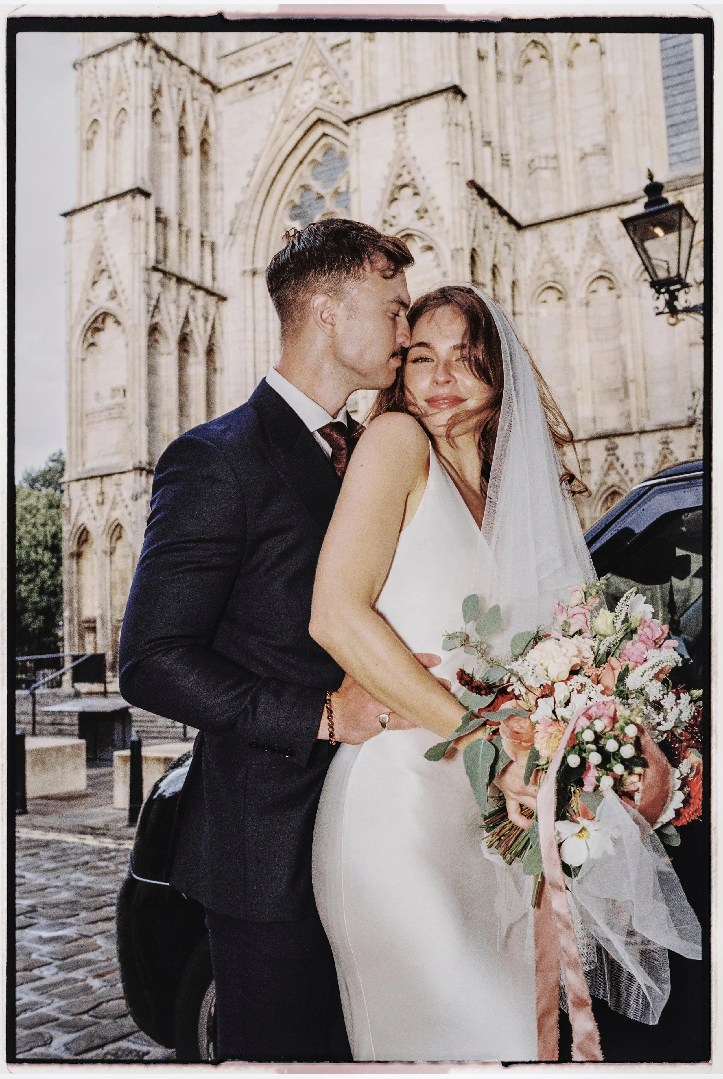 A newlywed couple embracing in front of a historic cathedral, with the groom kissing the bride's temple and the bride holding a bouquet of pink and white flowers, smiling softly.