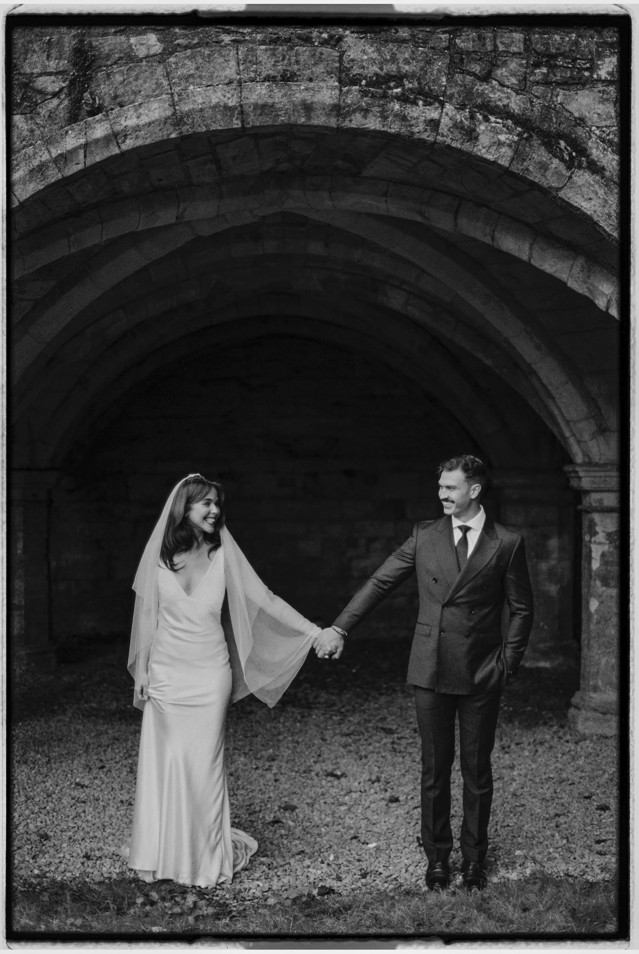 A bride and groom in wedding attire holding hands and smiling under an arched stone structure.