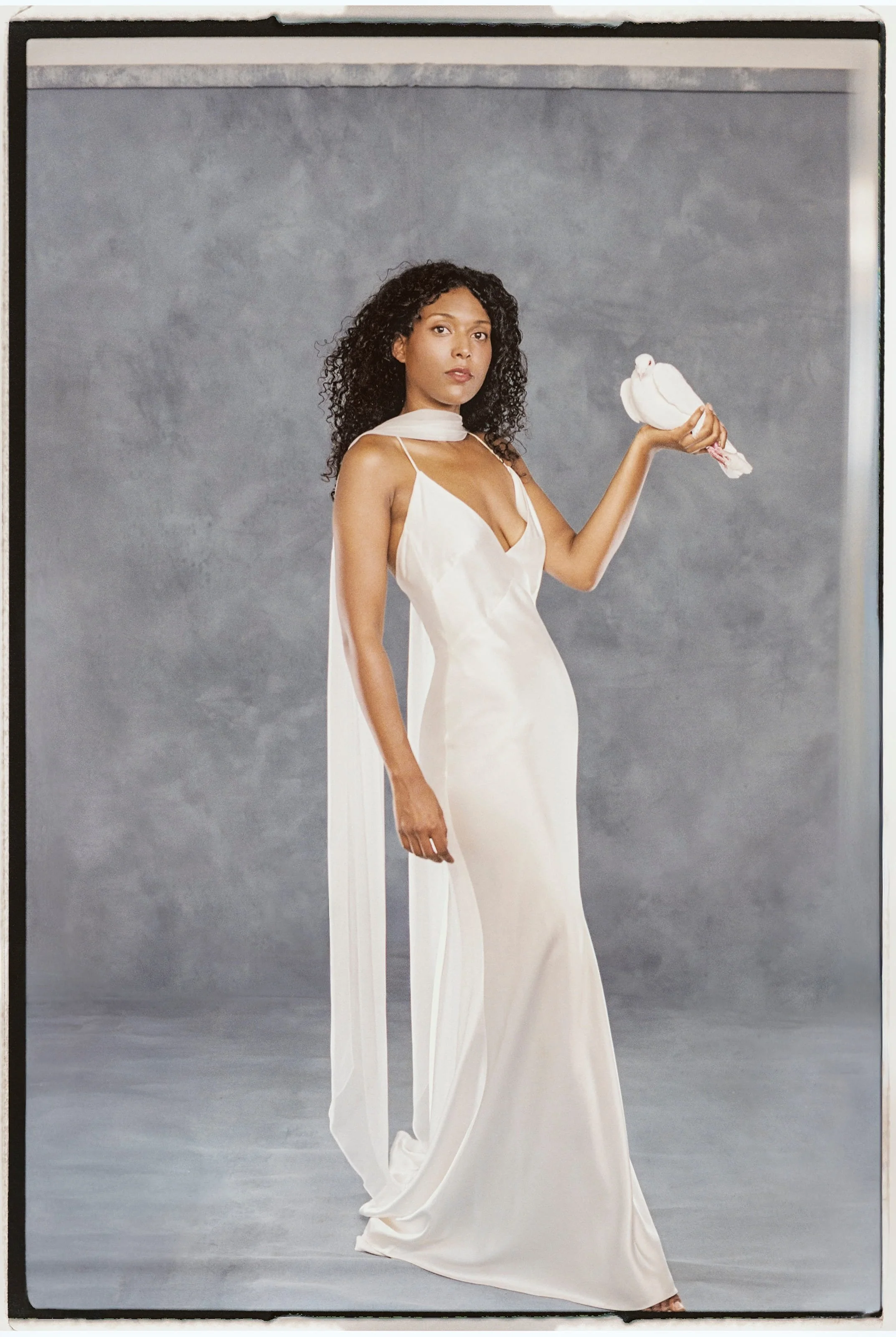 A woman with curly hair wearing a white satin gown with a deep neckline, holding a white dove against a gray backdrop.