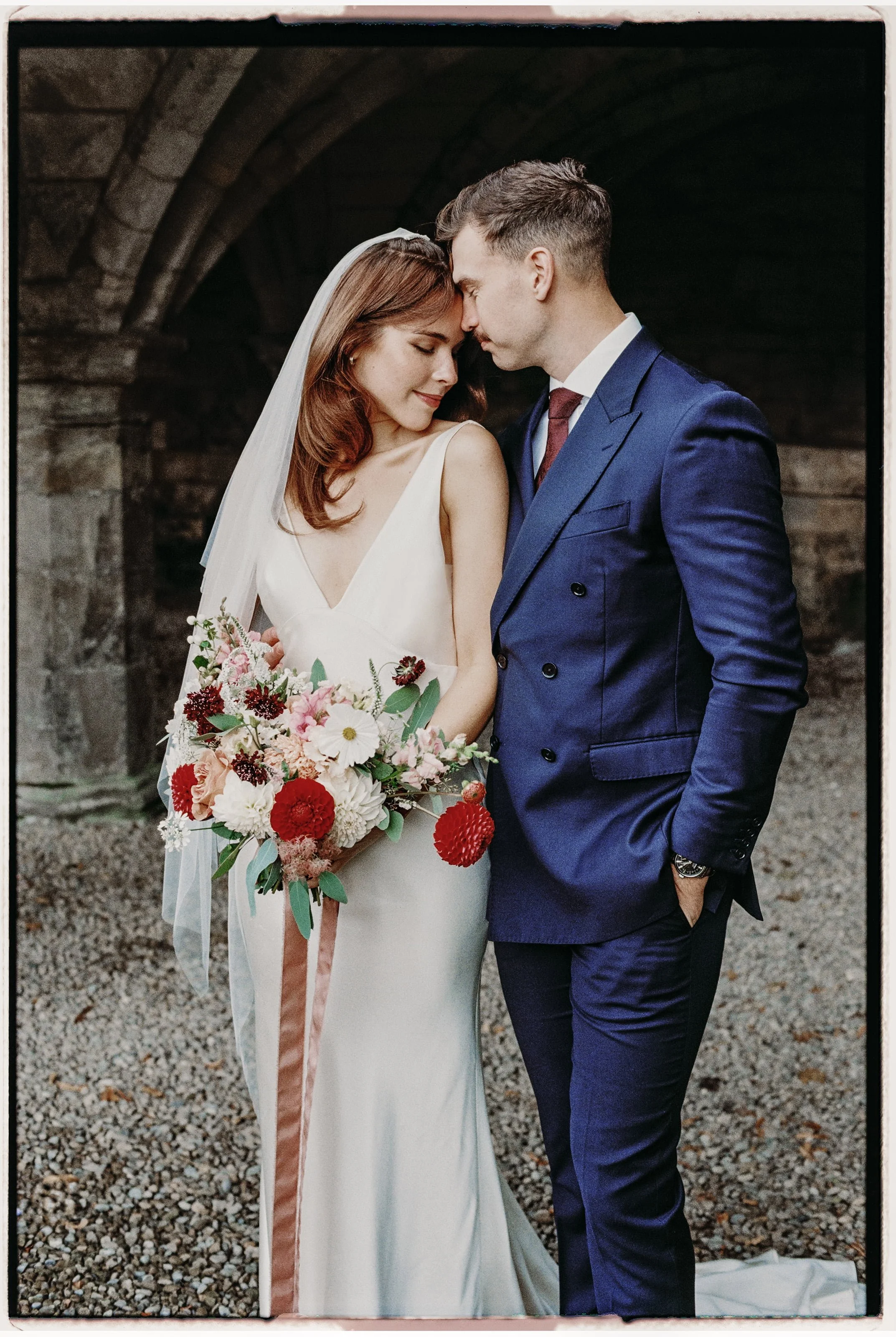 A bride and groom in wedding attire standing close with foreheads touching in front of a stone archway, the bride holding a bouquet of pink, white, and red flowers.