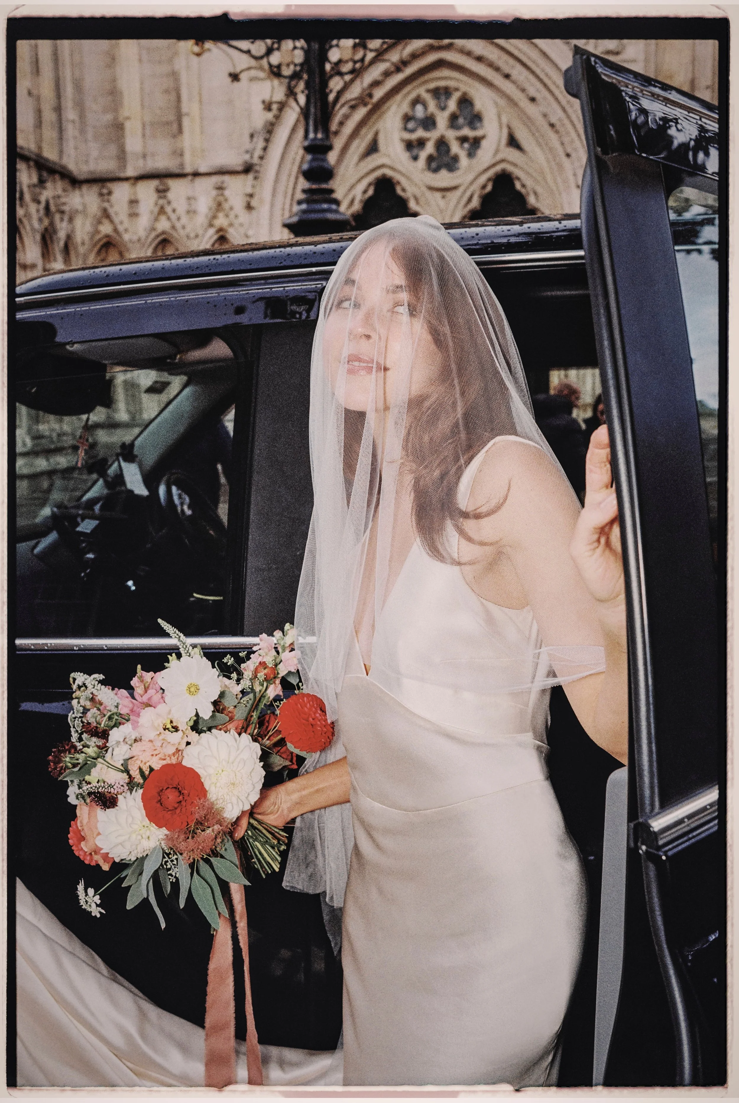 A bride in a satin wedding dress, holding a colorful bouquet, stands outside a church, partially inside a black vehicle, with a sheer veil over her face.