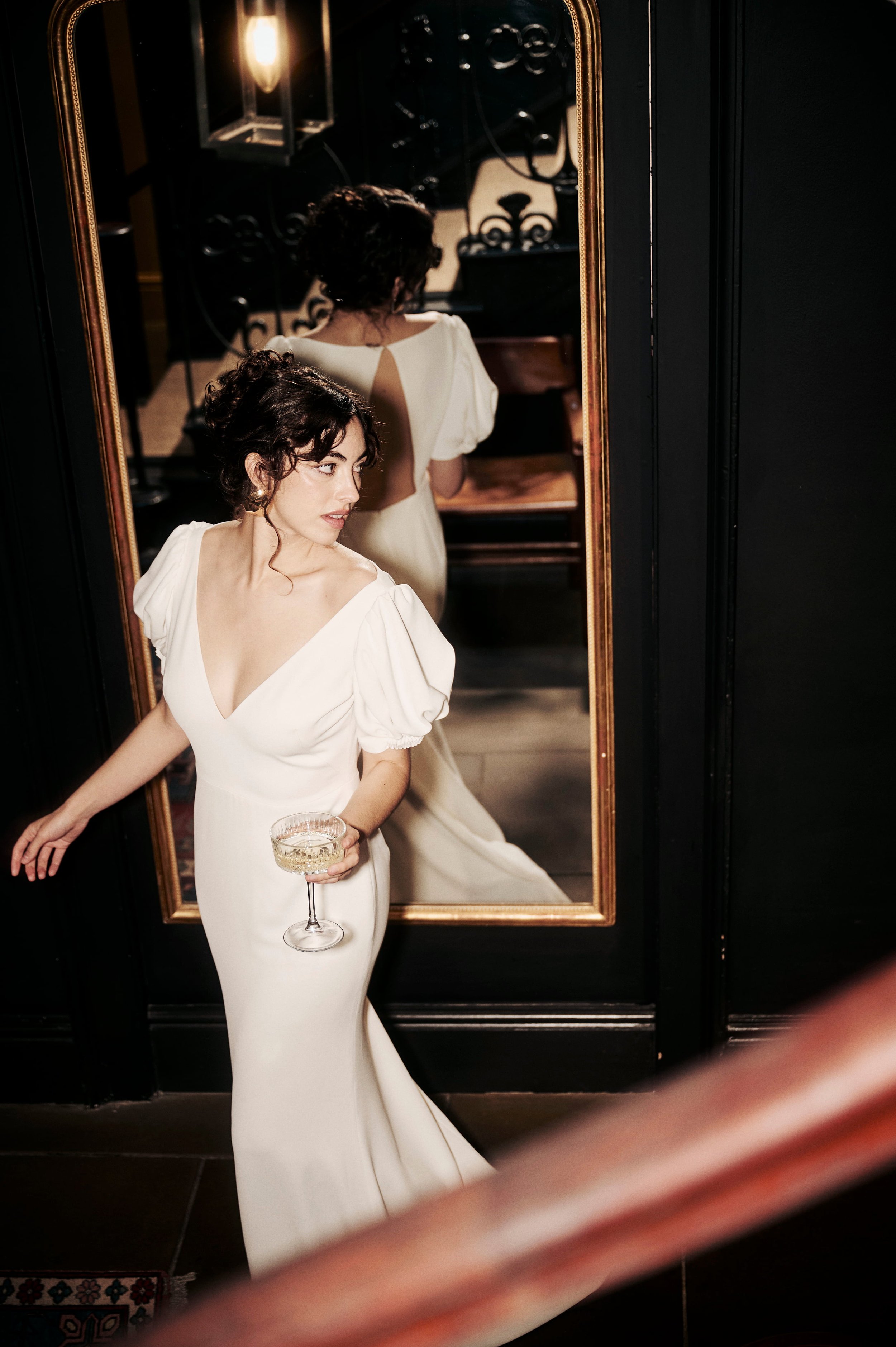 A woman in a white dress holding a glass of champagne, standing in front of a mirror, with her reflection showing her back, in a dimly lit room with dark walls and a wooden floor.