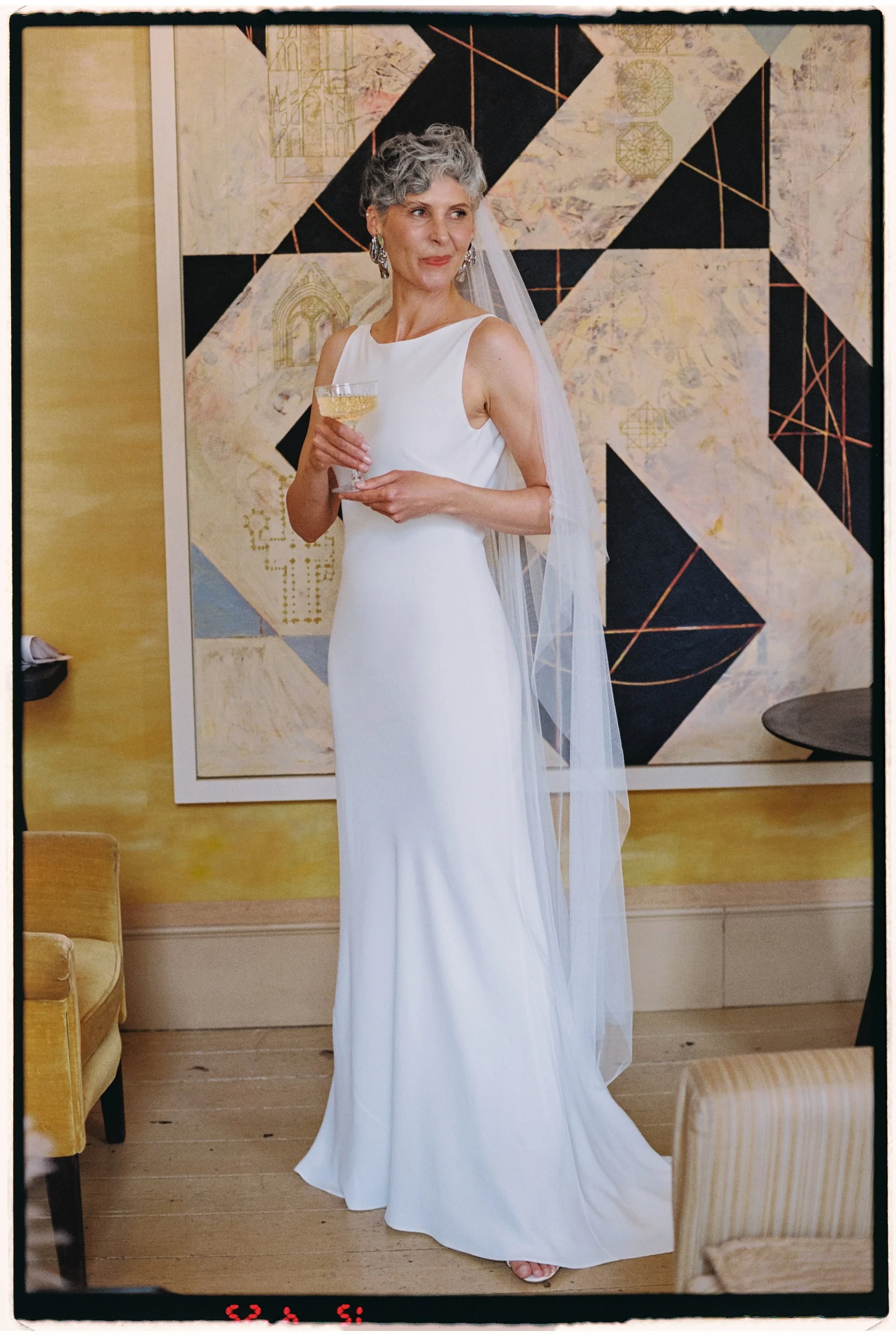 A mature woman with gray hair, wearing a white sleeveless wedding dress and veil, holding a glass of champagne, standing indoors in front of abstract artwork on the wall.