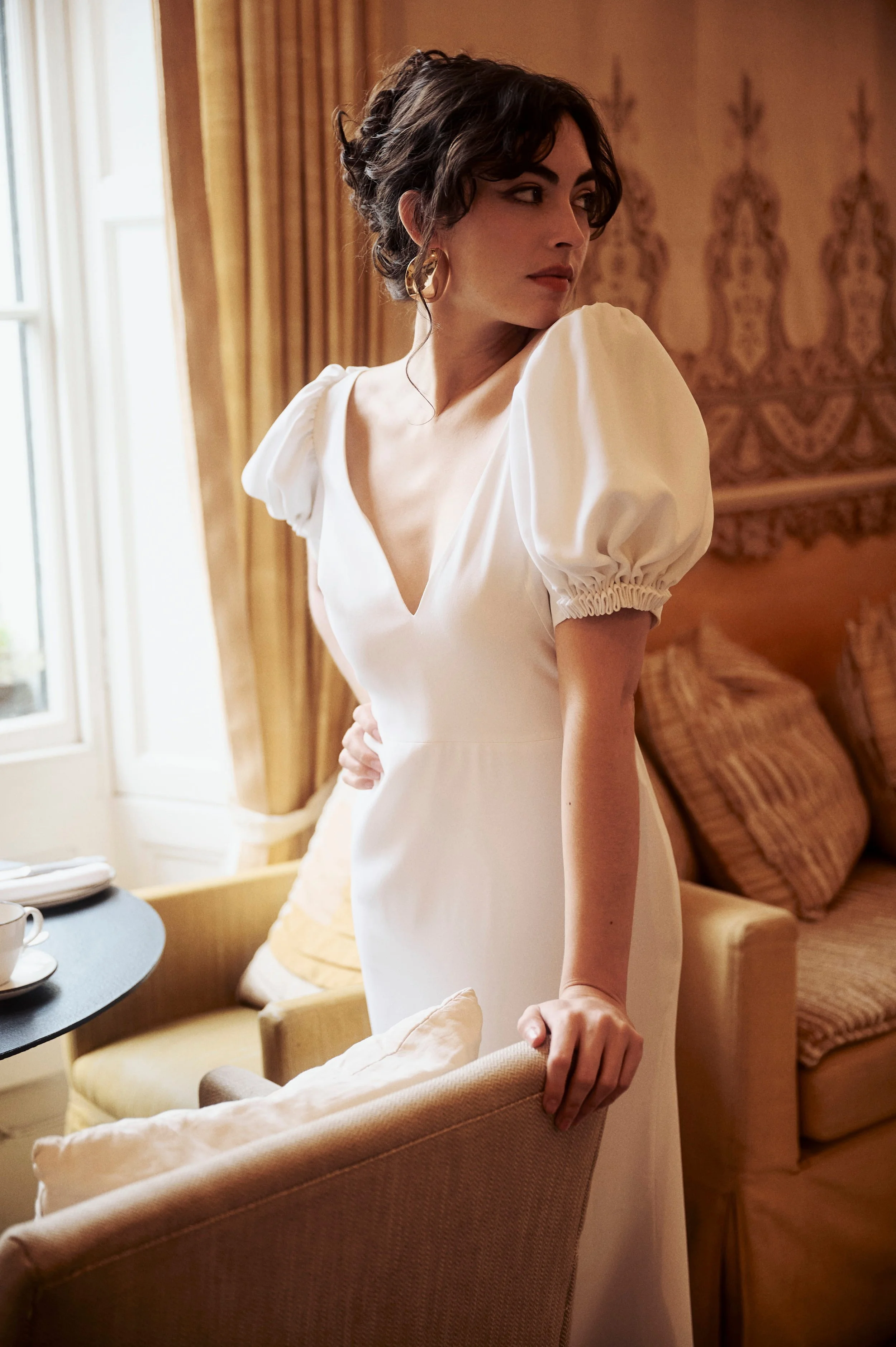 A woman in a white dress with puffed sleeves standing indoors near a window with golden curtains and wooden wall paneling, looking to her right.