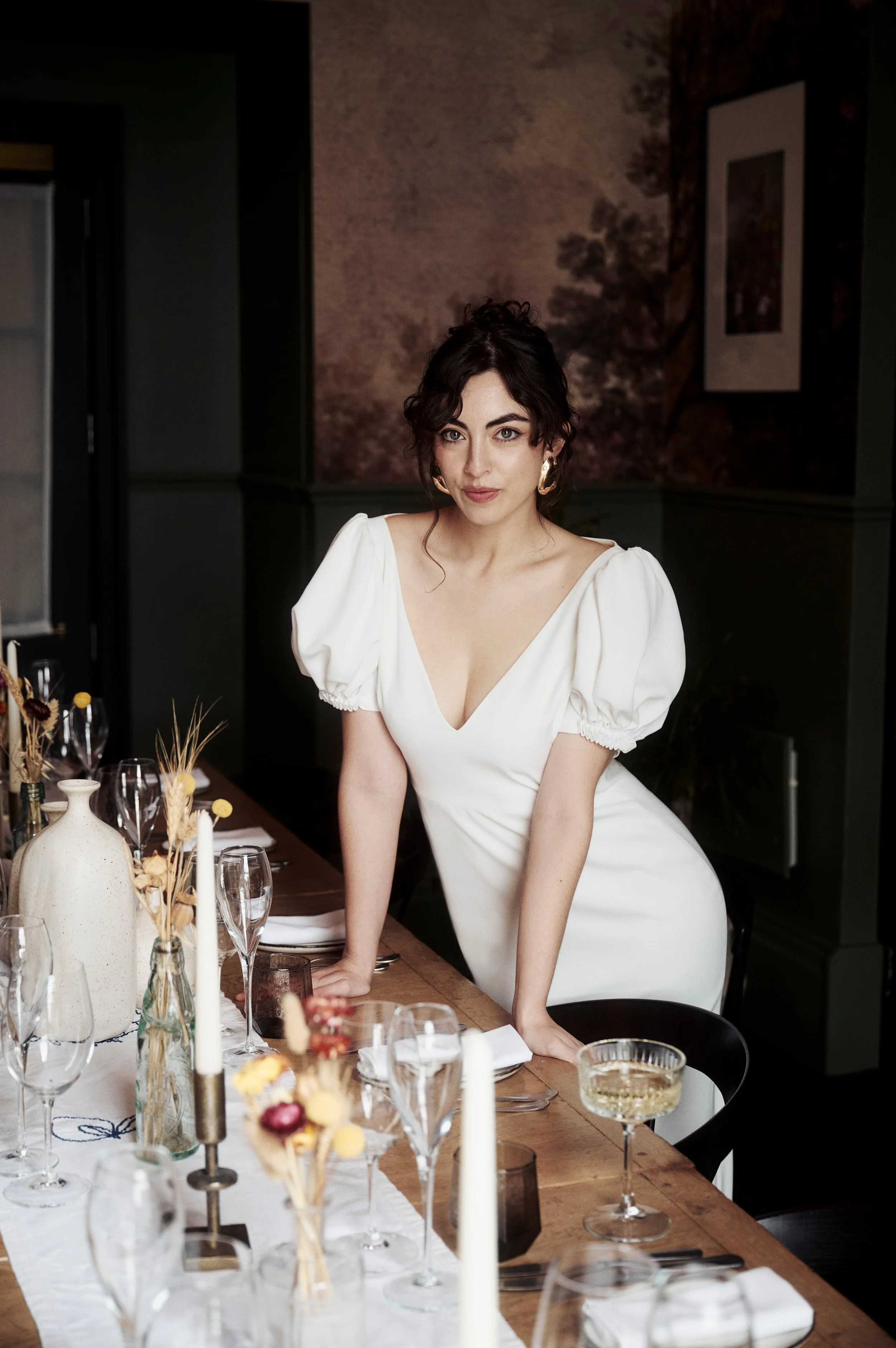 A woman with dark hair, wearing a white dress with puff sleeves, leaning on a dining table set with glassware and candles in a dimly lit room.