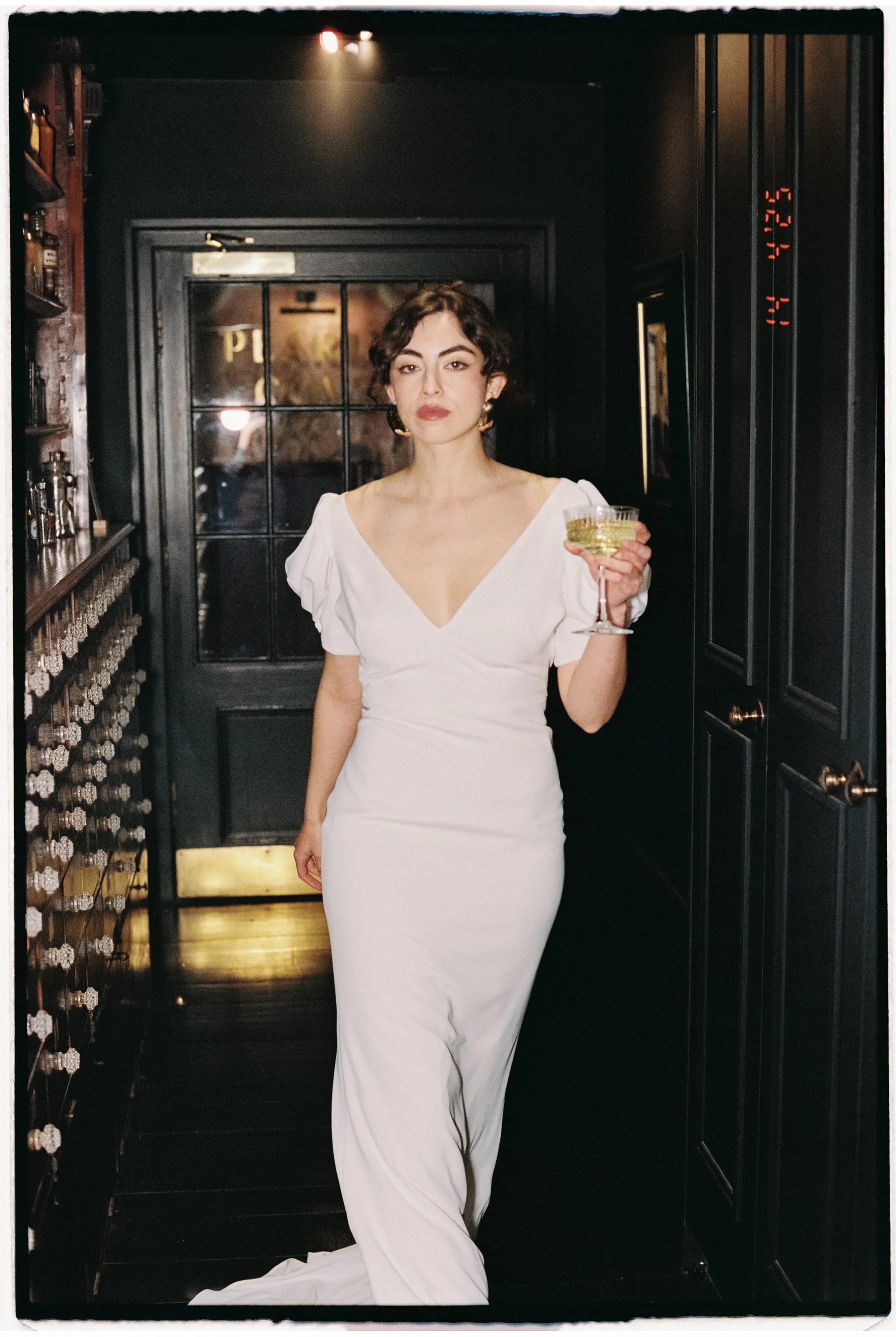 A woman in a white gown holding a cocktail glass, standing in a dimly lit bar or restaurant with dark walls and wooden shelves.