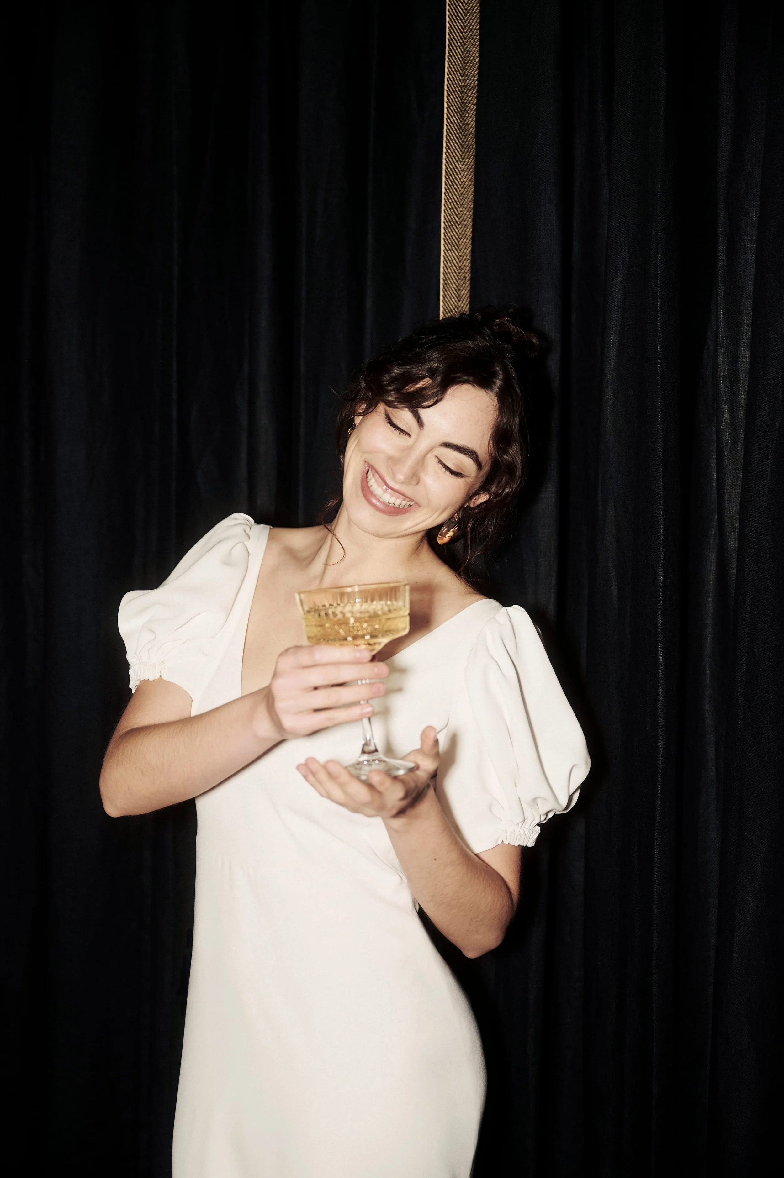 A woman with short curly brown hair wearing a white dress with puffed sleeves, holding a gold-colored cocktail glass, smiling and looking down.