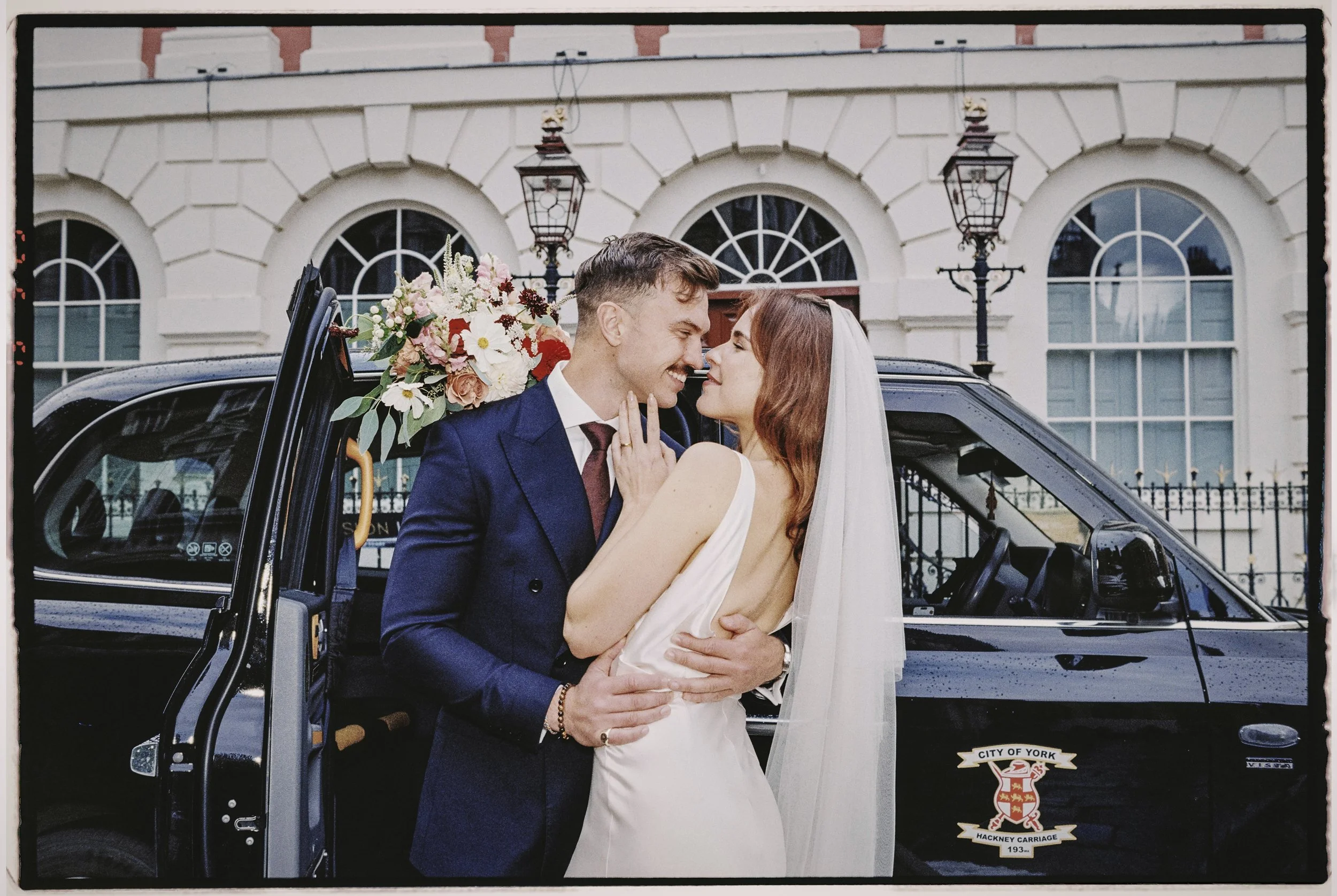 A newlywed couple is standing close together in front of a black vehicle with a bouquet of flowers in the background. The groom is wearing a dark blue suit, and the bride is wearing a white wedding dress and veil. They are gazing into each other's eyes, smiling, in front of a large building with arched windows and vintage street lamps.