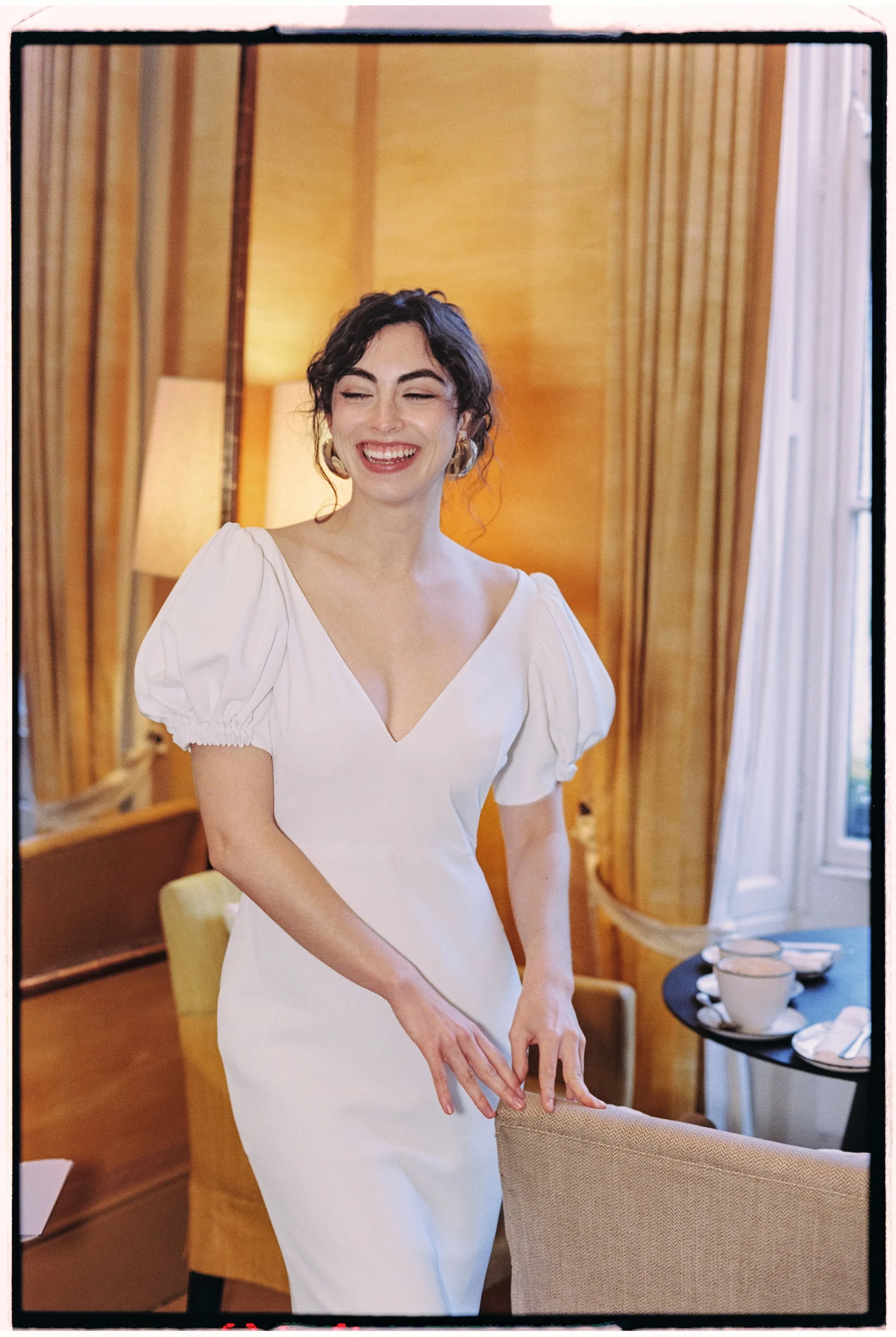 A woman in a white dress with puffed sleeves, smiling and standing in a warmly lit room with wooden paneling, curtains, and a window, with a table set with cups and plates in the background.