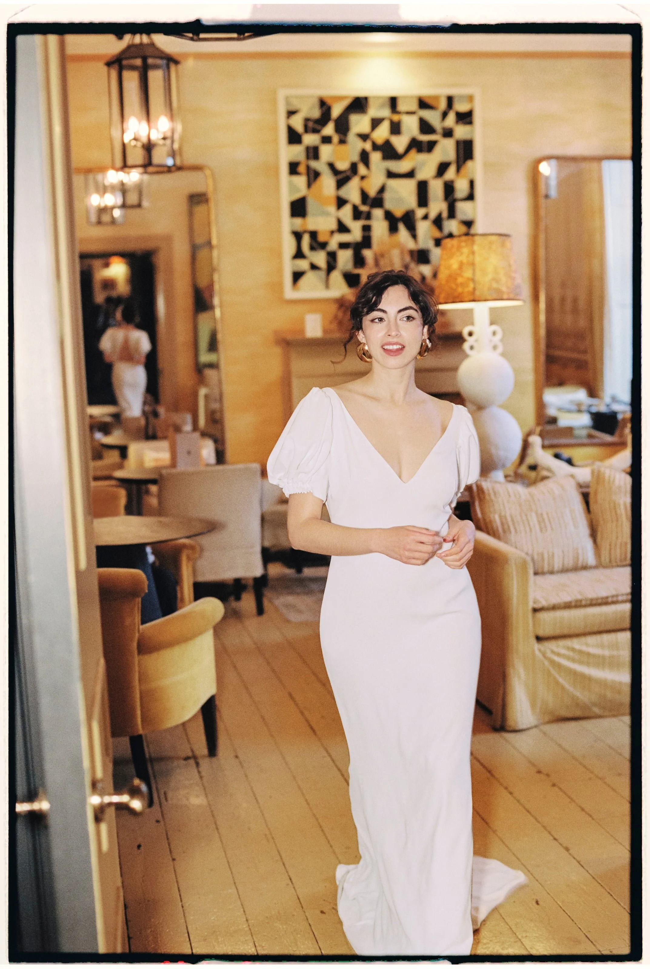 A woman in a white dress standing inside a warmly lit room with wooden floors, armchairs, a table, and decorative lamps.