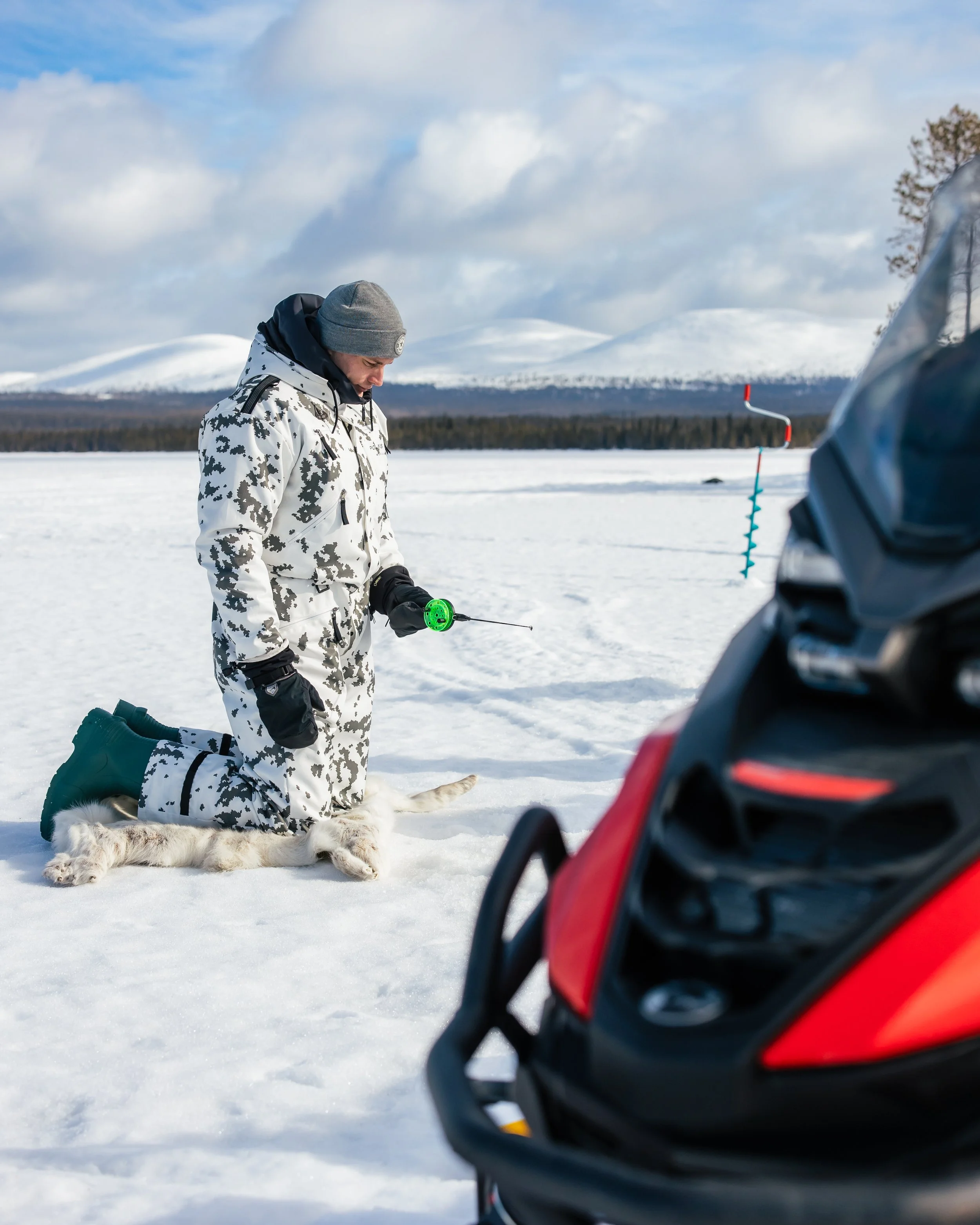A person kneeling on snow, dressed in camouflage winter gear, holding a fishing rod, with an all-white dog lying beside them, in a snowy landscape with mountains in the background. A snowmobile is partially visible in the foreground.