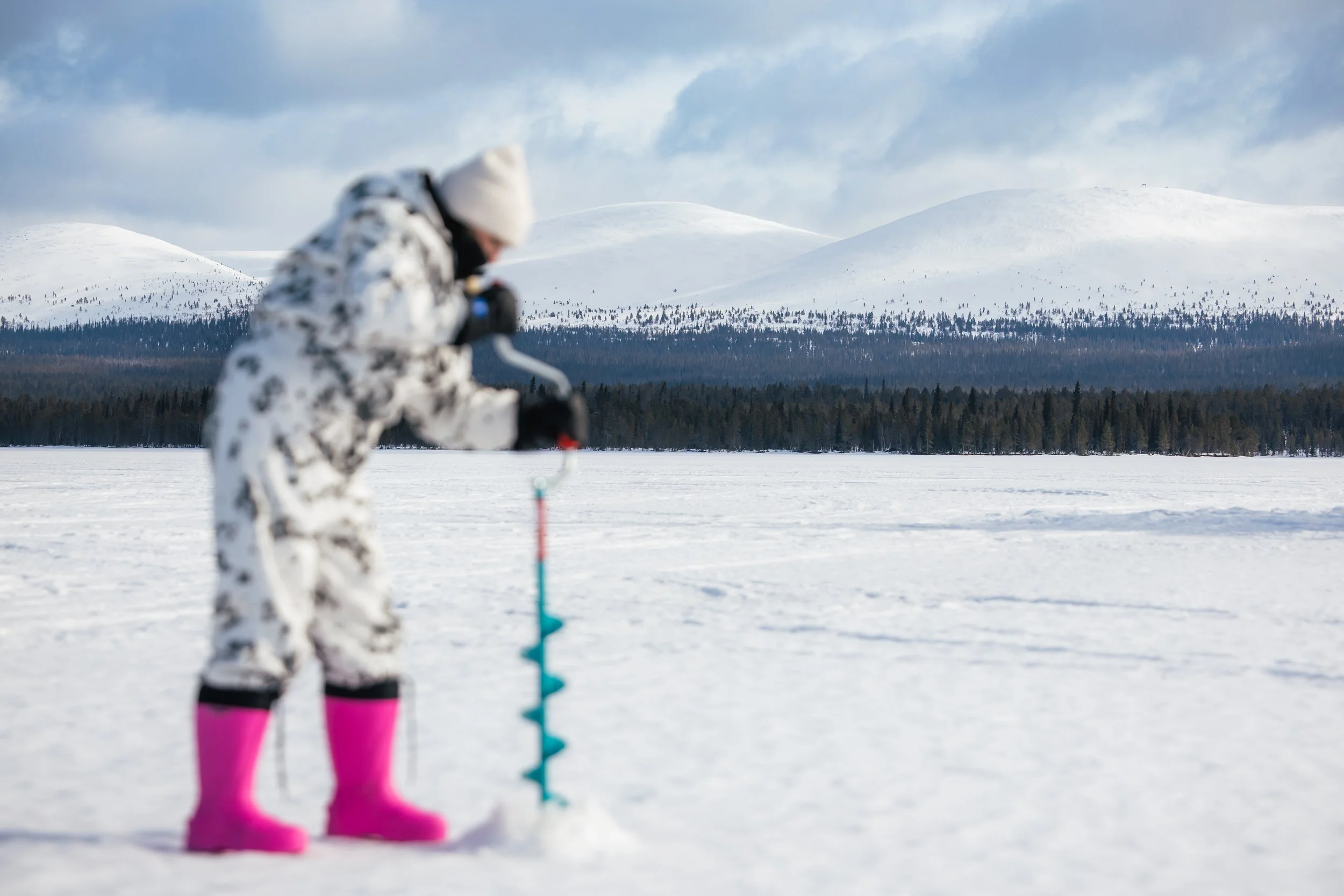 Child dressed in white camouflage winter suit with pink boots, using an ice auger on a frozen lake with snow-covered mountains and a forest in the background.