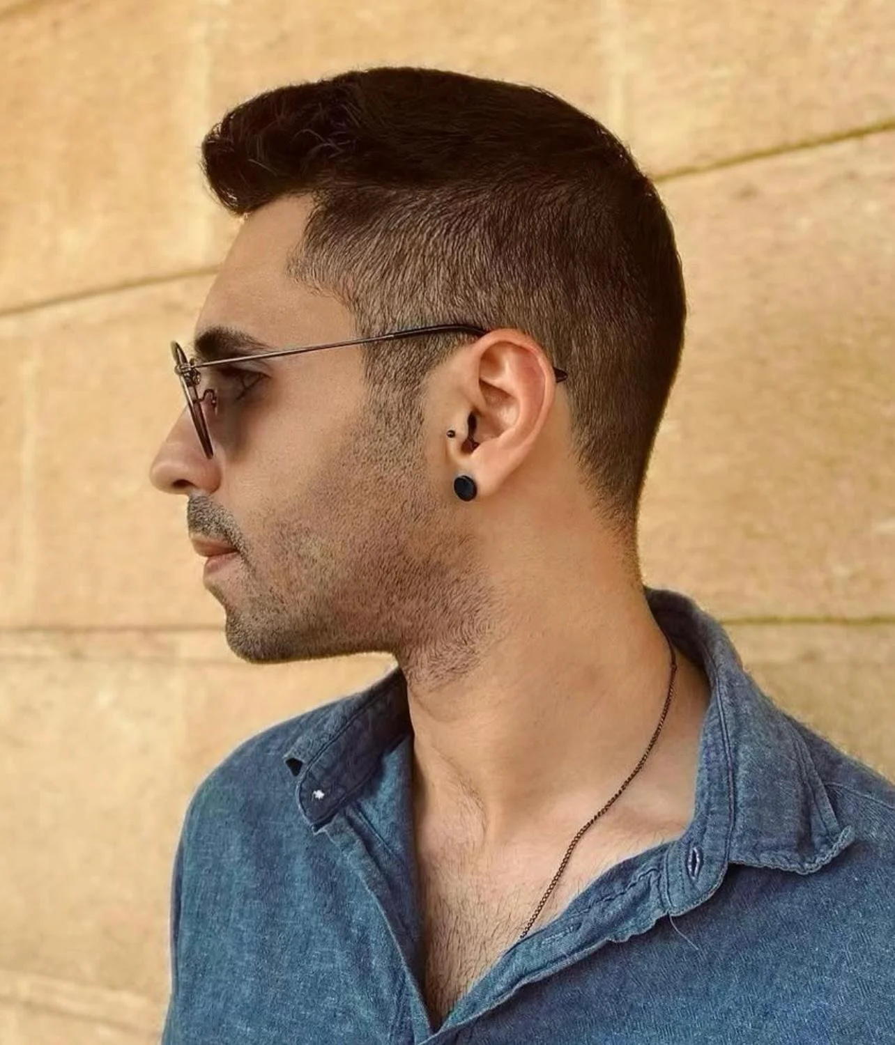 Side profile of a man with dark hair, sunglasses, and multiple earrings, wearing a denim shirt, against a tan wall.