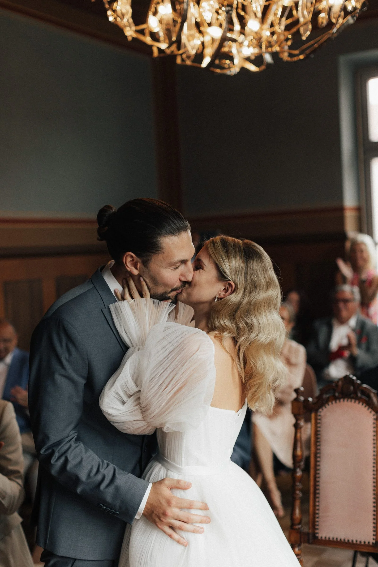 Ein Brautpaar küsst sich bei ihrer Hochzeit in einem festlichen Raum voller Gäste mit einem Kronleuchter an der Decke.