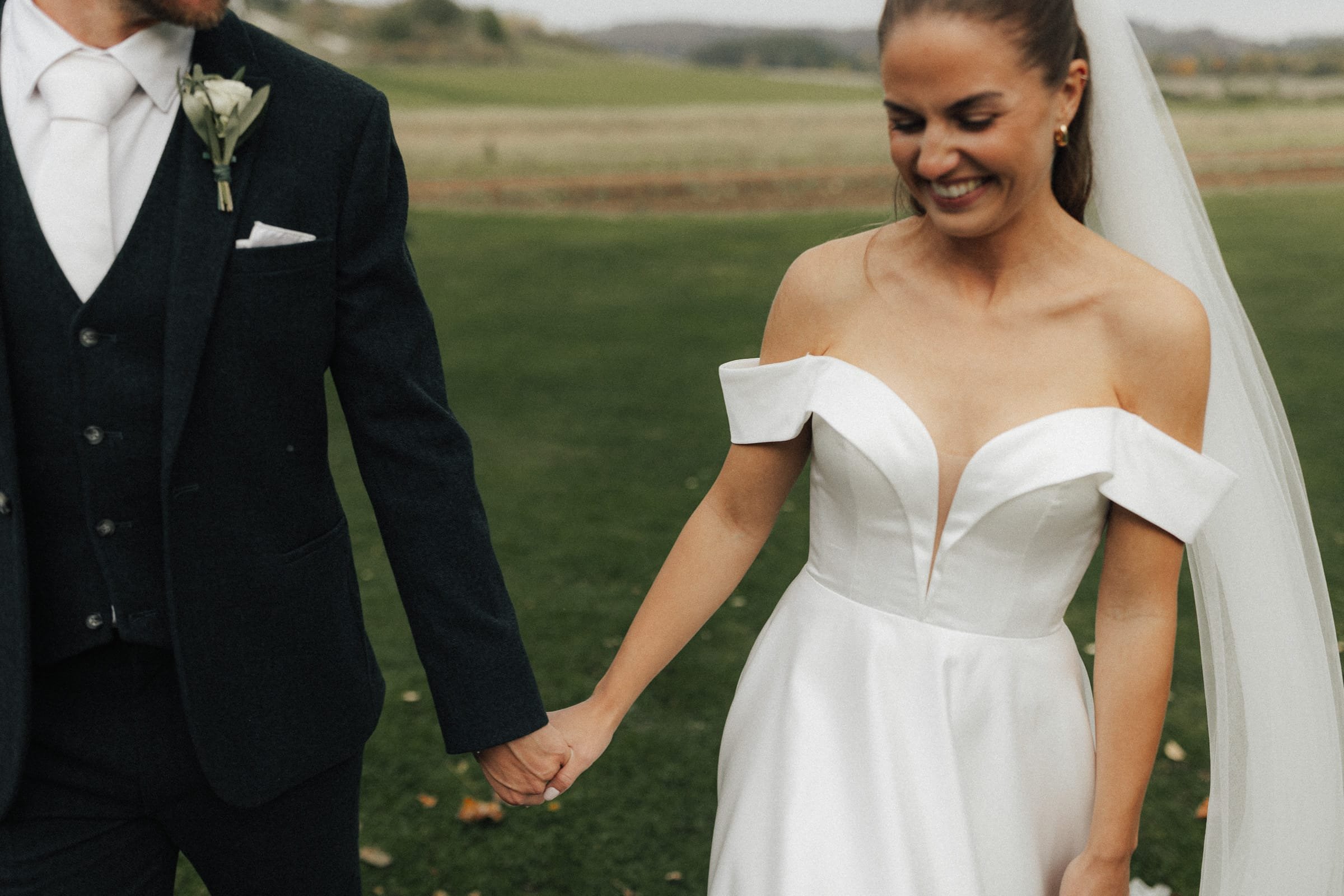 Ein Brautpaar hält Händchen im Freien, das Kleid der Braut ist weiß mit schulterfreien Details, der Bräutigam trägt einen schwarzen Anzug mit weißem Hemd und Krawatte.