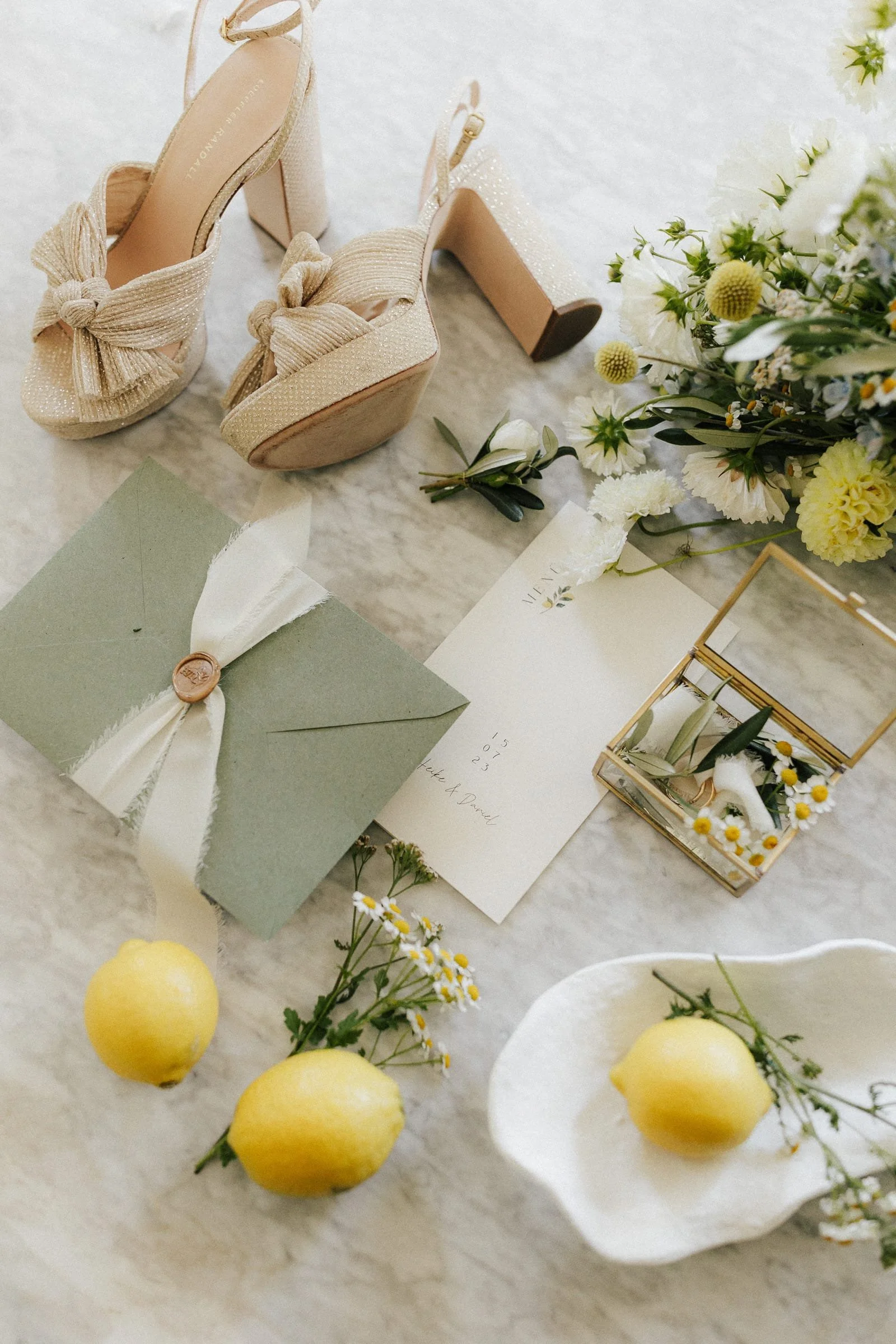 Elegante goldene High Heels von Loeffler Randall mit Schleifen, eine grüne Einladungskarte, eine weiße Karte mit Blumen, eine kleine Schmuckbox mit Blumen, drei Zitronen, weiße Blumen und ein Blumenstrauß.