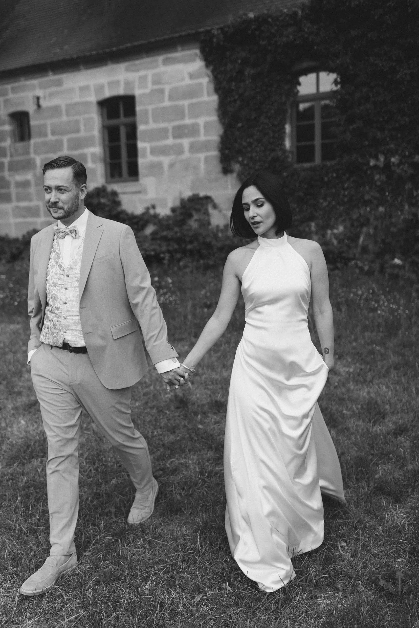 Braut und Bräutigam halten Händchen beim Spaziergang auf einer Wiese, im Schwarz-Weiß-Foto.