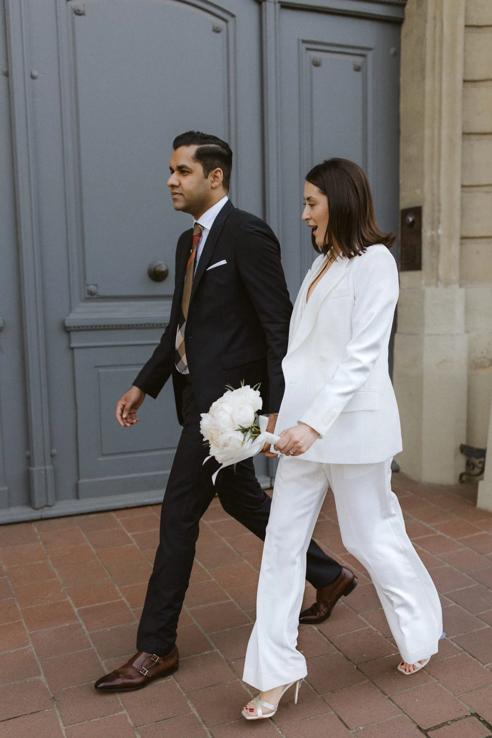 Ein romantisches Paar im Anzug und Hochzeitskleid geht Hand in Hand vor einer großen, grauen Tür.