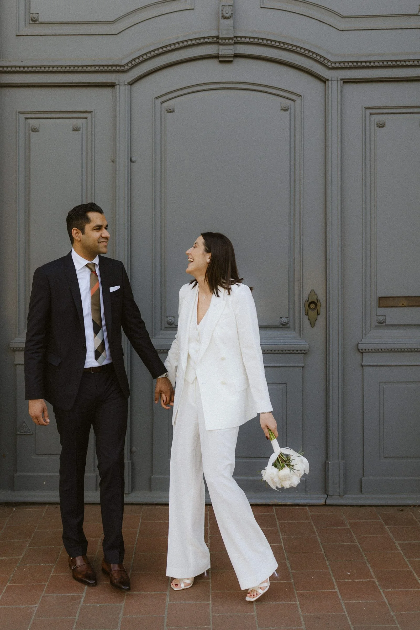 Ein Mann und eine Frau in formeller Kleidung, die Händchen halten und lachen, vor einer grauen Doppeltür, die Frau trägt einen weißen Anzug und hält einen weißen Blumenstrauß.