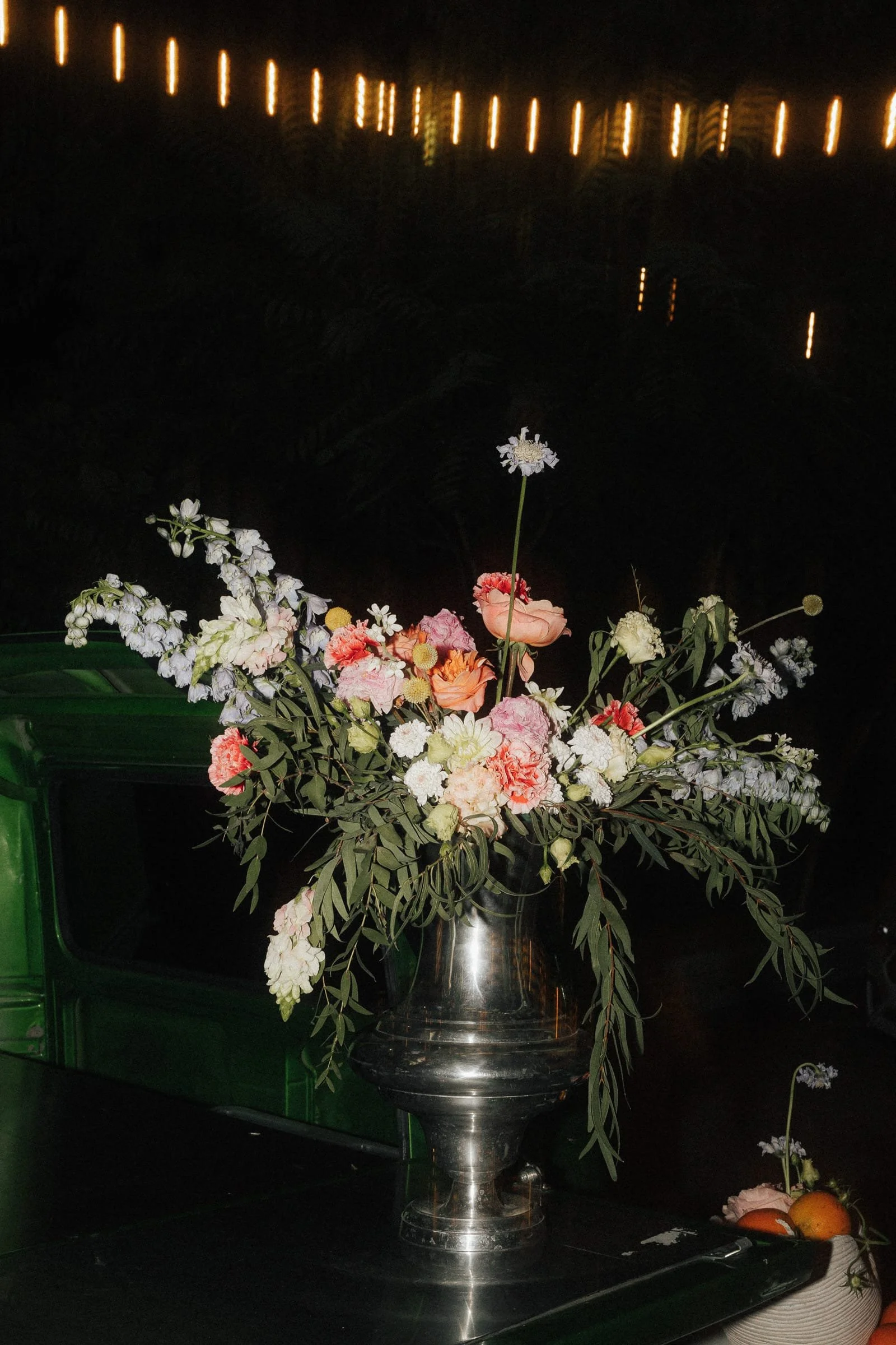 Blumenstrauß in einer silbernen Vase auf einem schwarzen Tisch, dunkler Hintergrund mit Lichtern.