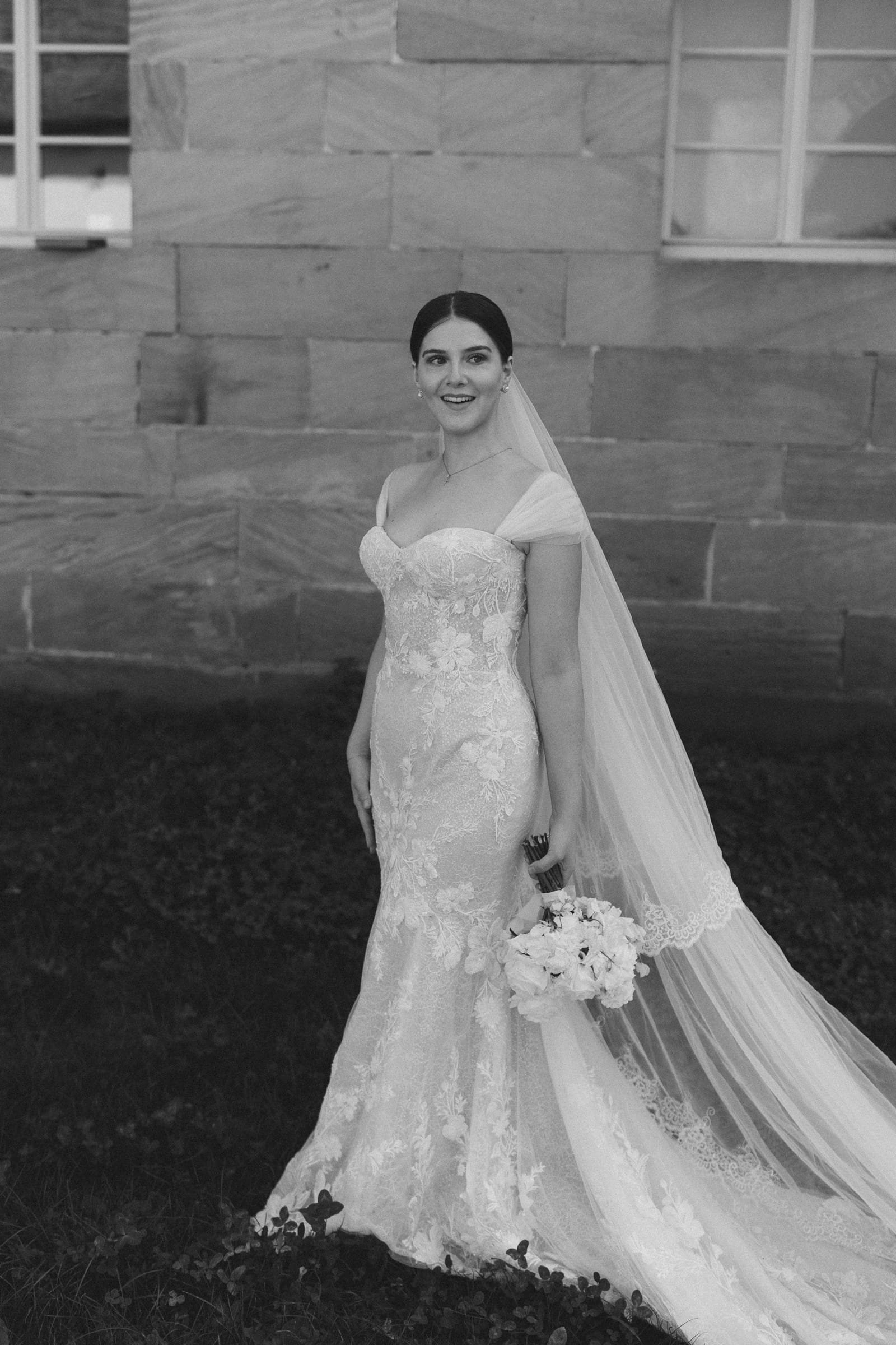 Eine Braut in einem eleganten, weißen, makellosen Hochzeitskleid mit Spitzenverzierungen, die lächelnd vor einer Steinmauer steht, während sie eine Hand mit einem Blumenstrauß hält.