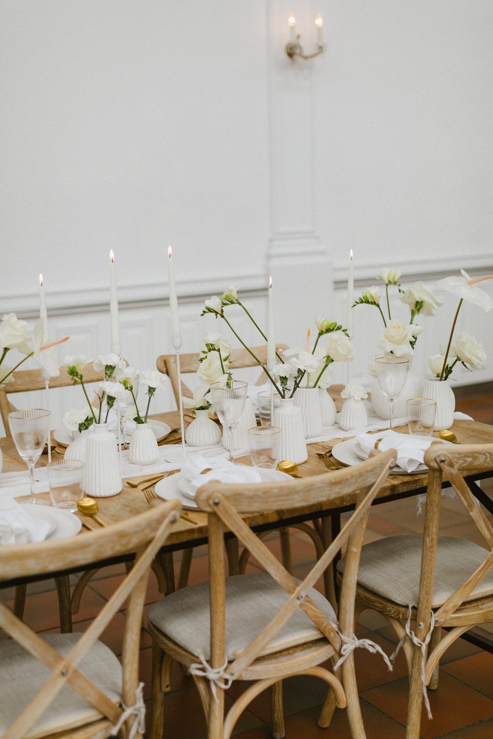 Ein festlich gedeckter Tisch mit weißen Vasen, weißen Blumen, Kerzen und goldenen und weißen Geschirrplatten in einem eleganten Raum.