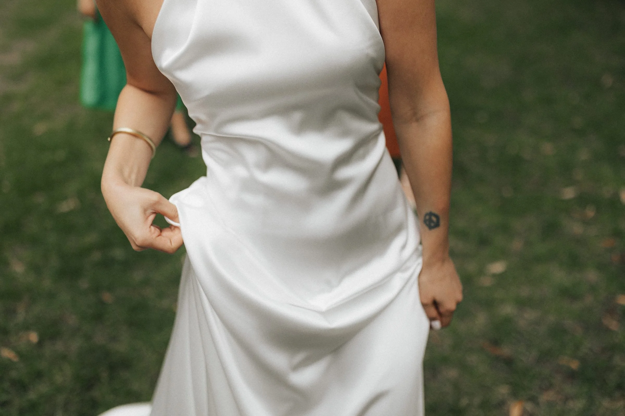 Nahaufnahme eines weißen Kleides, getragen von einer Person, die auf einem grasbewachsenen Außenbereich steht, mit sichtbarer Tattoo am linken Unterarm.