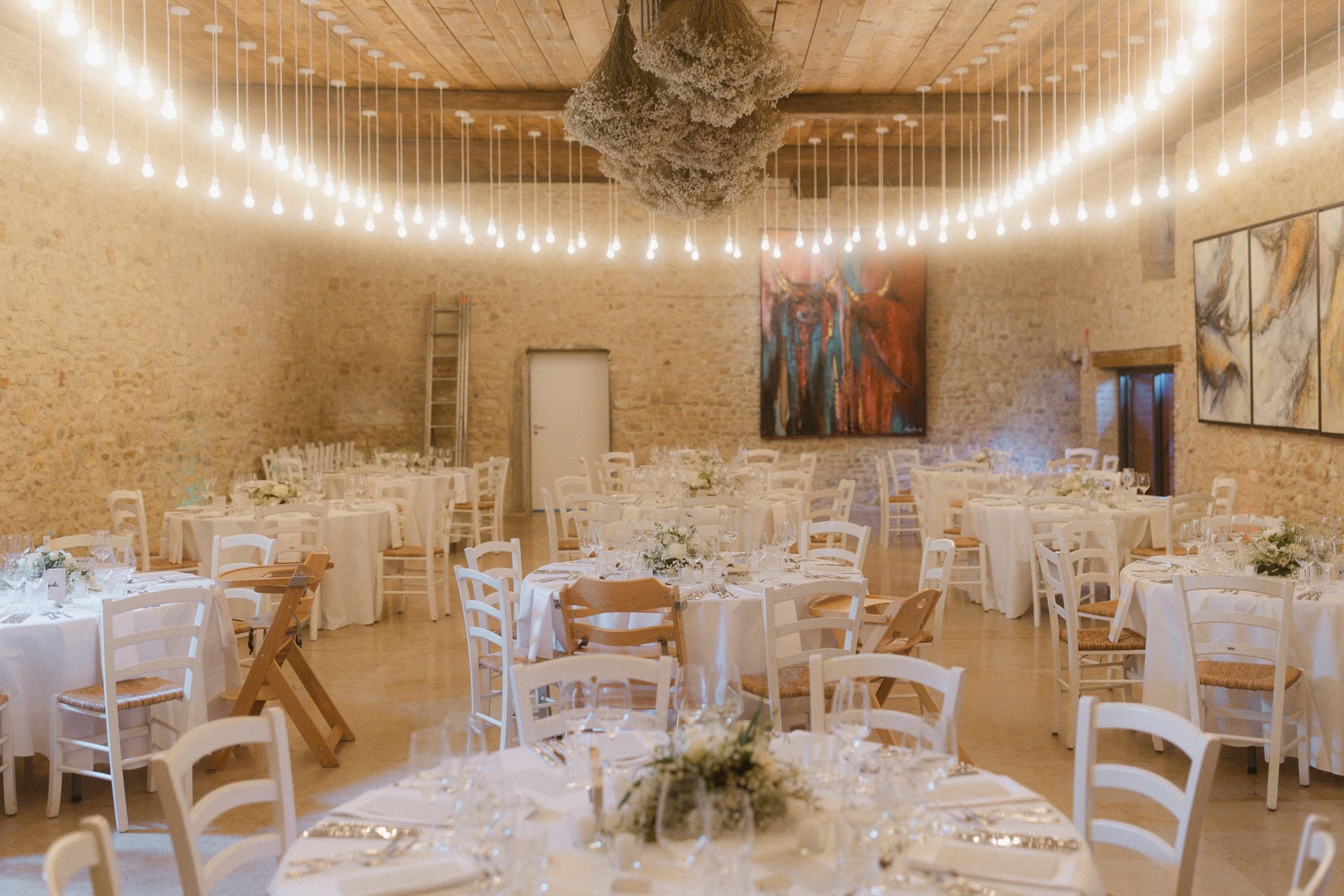 Ein festlich gedeckter Saal mit weißen Tischen, sitzenden Stühlen, Blumenarrangements, warmen Lichterketten an der Decke und Wandkunst, vorbereitet für eine Feier.