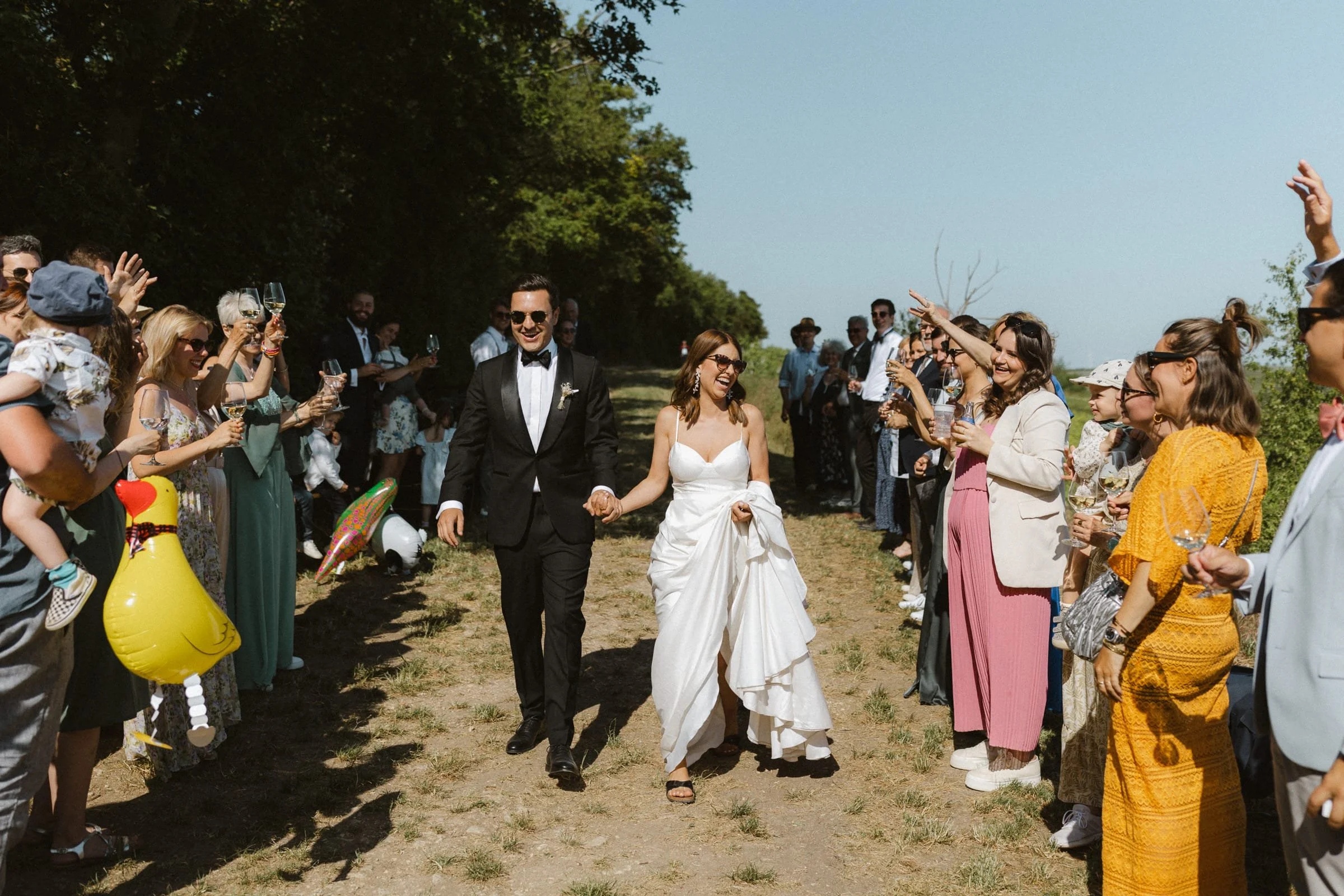 Ein lachendes Brautpaar geht Hand in Hand durch eine Menschenmenge, die auf beiden Seiten stehen und mit Gläsern anstoßen, während sie draußen an einem sonnigen Tag feiern.