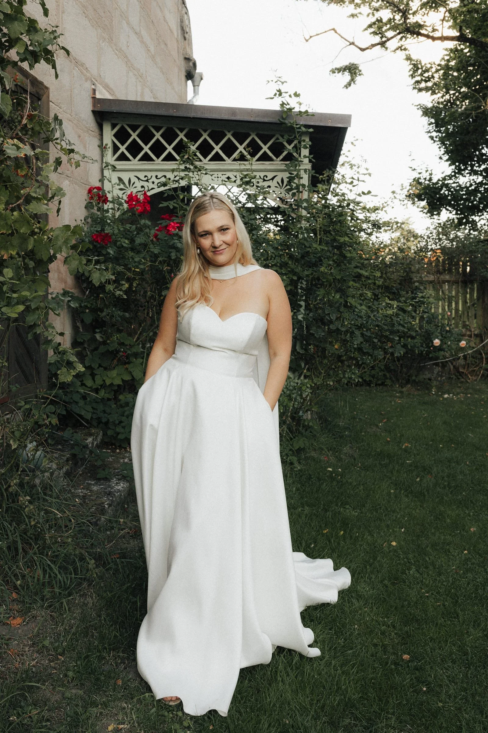 Eine Frau in einem weißen Brautkleid steht im Garten, umgeben von Blumen und Büschen, bei Tageslicht.