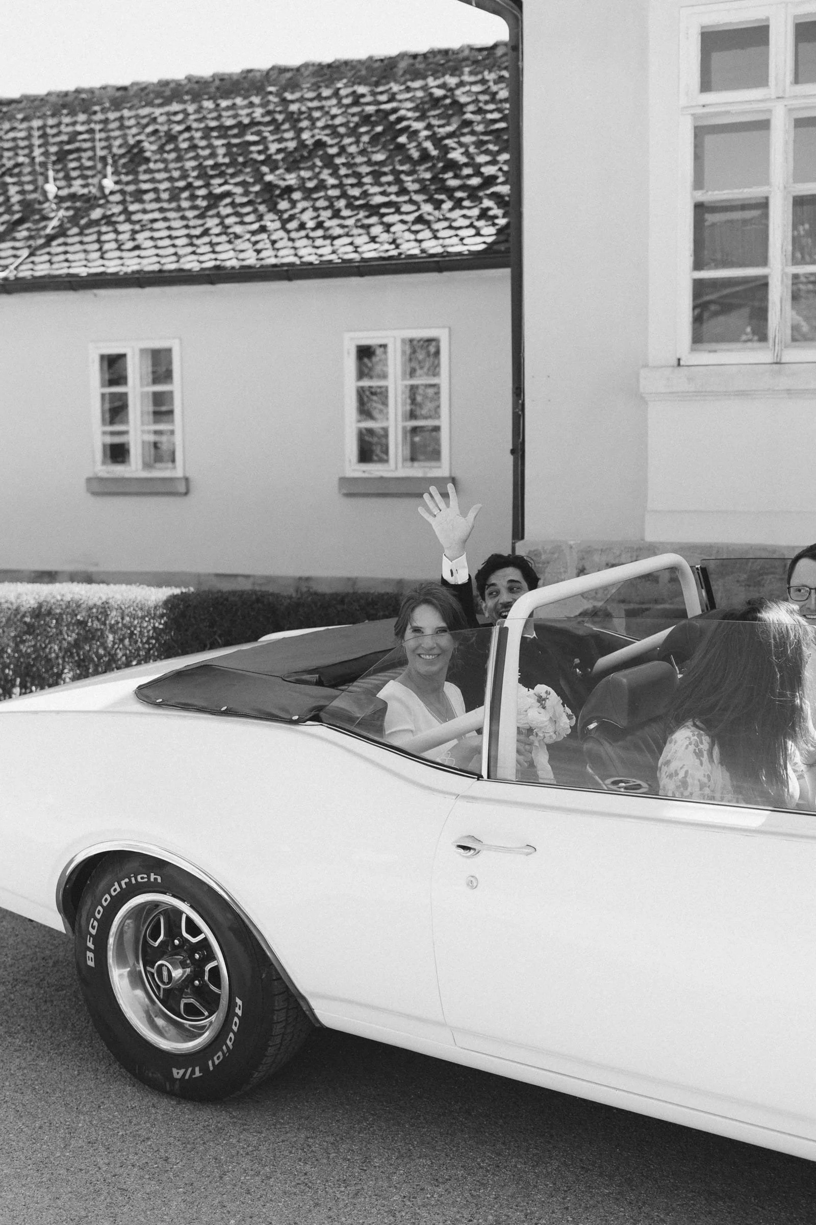 Braut und Bräutigam sitzen in einem weißen Cabrio, die Braut hält einen Blumenstrauß, der Bräutigam winkt, alle lachen, im Hintergrund ein Haus mit Fenstern, das Bild ist in Schwarz-Weiß.