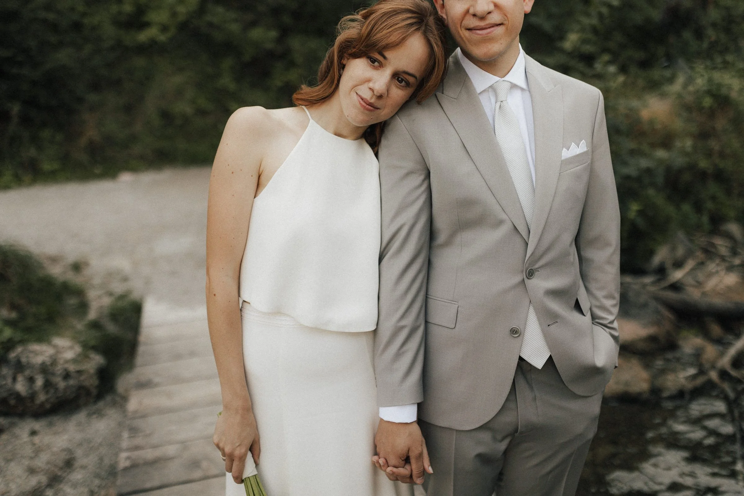 Ein hochzeitsjunges Paar, das sich an einem Steg am Wasser hält, mit Händen zusammen, in formeller Kleidung, umgeben von Natur.