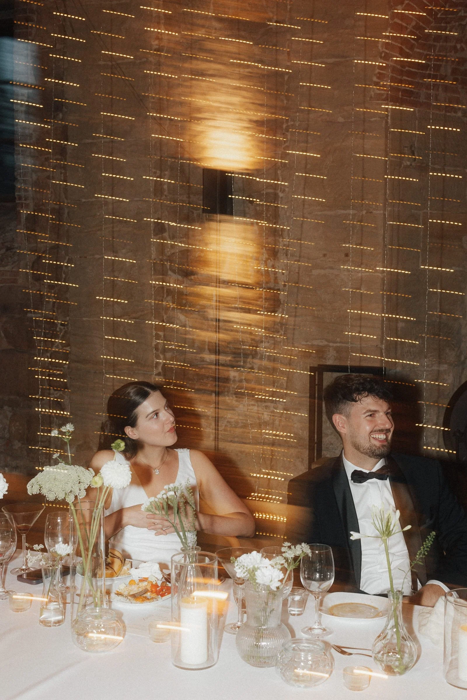 Ein Brautpaar bei einer Hochzeitsfeier, sitzend an einem festlich gedeckten Tisch mit Blumen und Kerzen, im Hintergrund Lichter an einer Steinwand.